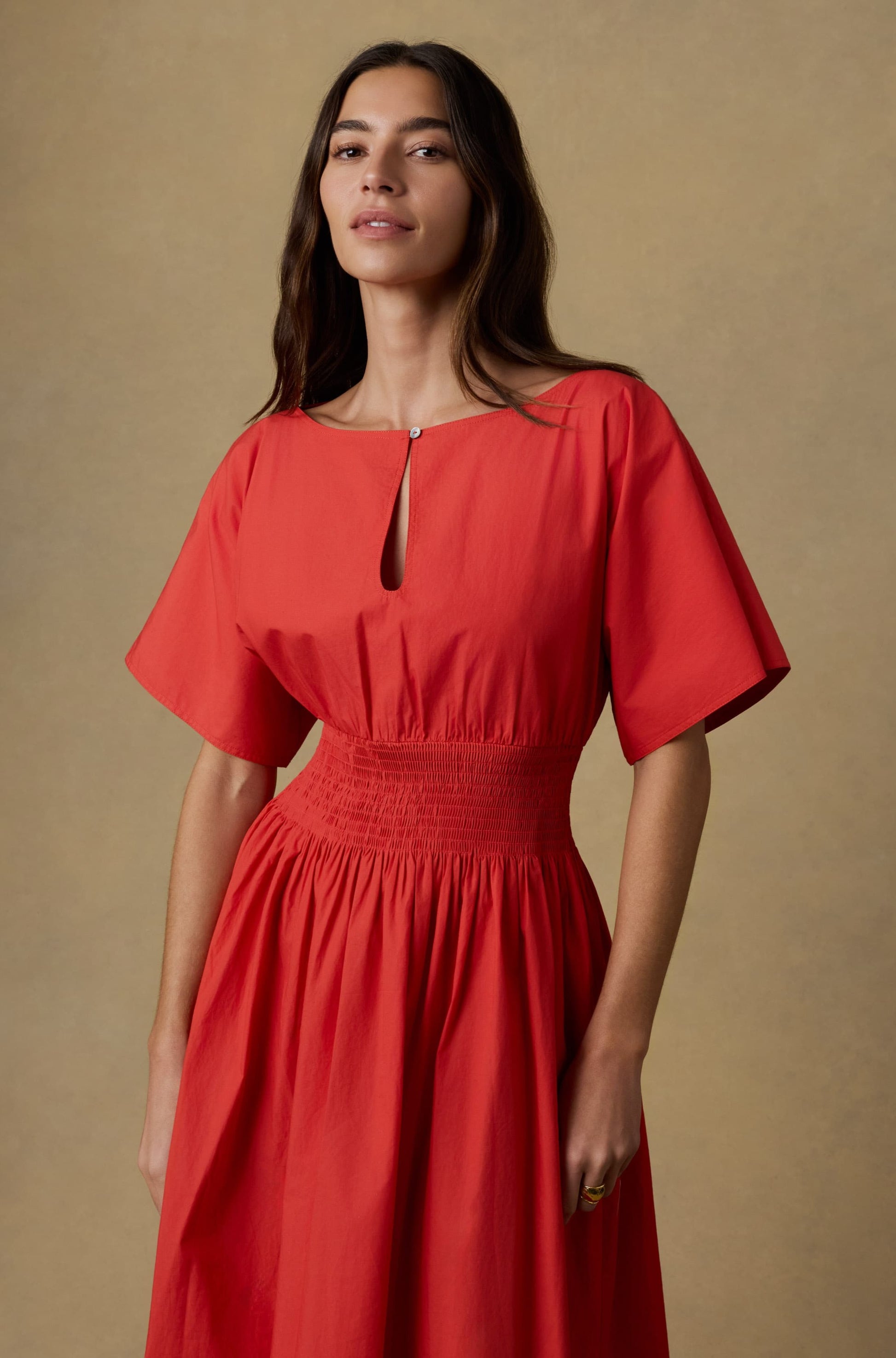 A woman with long brown hair wears the Faherty Coastal Poplin Elia Smocked Dress, a red maxi with short sleeves and a gathered waist, standing against a plain beige background.