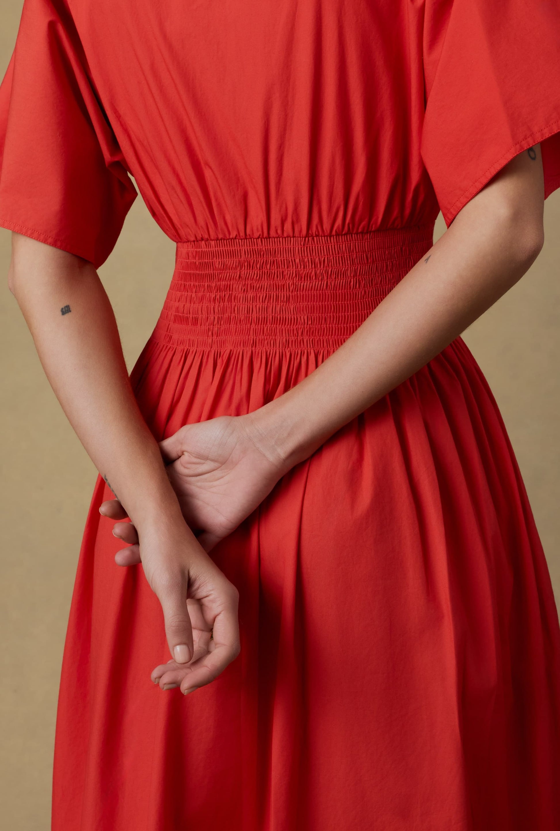 A person in a red Coastal Poplin Elia Smocked Dress by Faherty stands with their back to the camera, hands clasped behind them. The A-line cotton dress features shirring at the waist. A small square tattoo is visible on their left forearm, against a beige background.