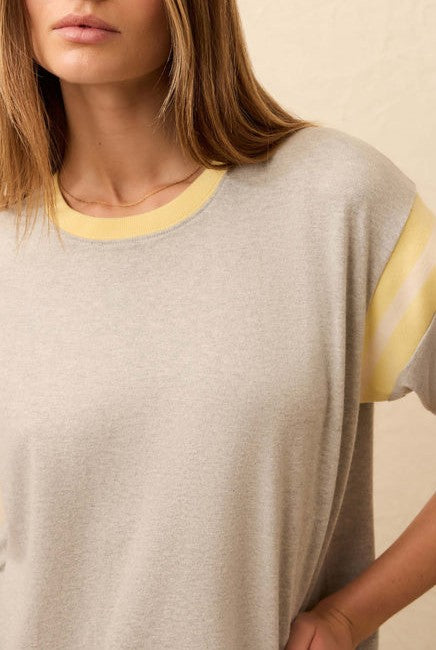 A woman with straight, light brown hair models the Faherty Sport Jersey Throwback Tee, a retro-inspired light gray short-sleeve top with yellow and white sleeve stripes and a yellow-trimmed neckline, posed against a beige background.