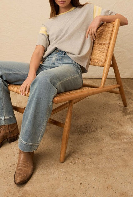 A person sits sideways on a woven wooden chair, wearing the Faherty Sport Jersey Throwback Tee in light gray with faded blue jeans and brown leather boots. The photo is cropped above the shoulders; the floor is bare concrete.