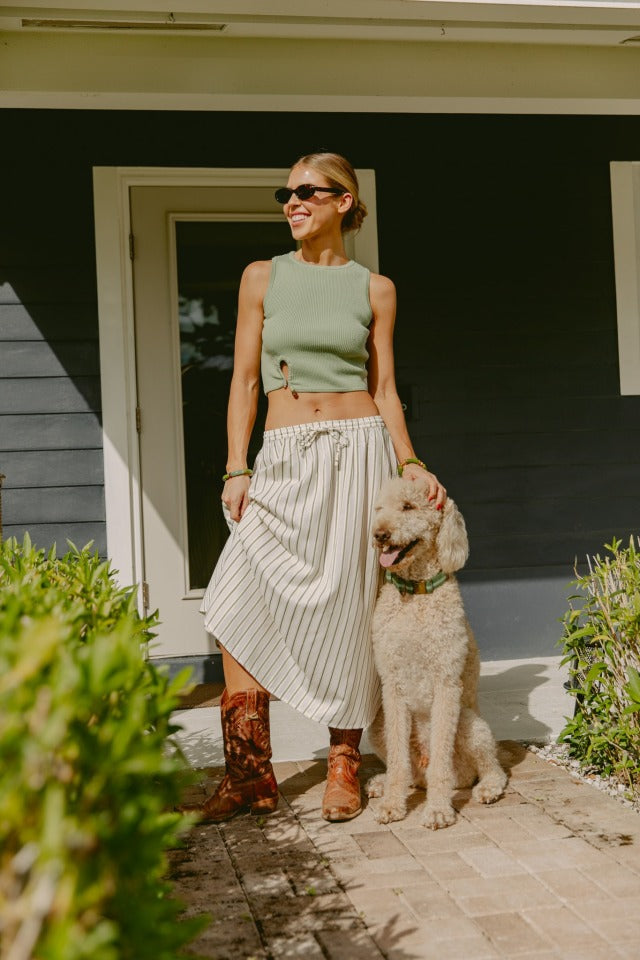 Wearing the Isla x Tate Tyler Hardware Sweater Tank, a woman in sunglasses, a striped white skirt, and brown boots smiles outside a house, resting her hand on a large curly-haired dog sitting beside her.