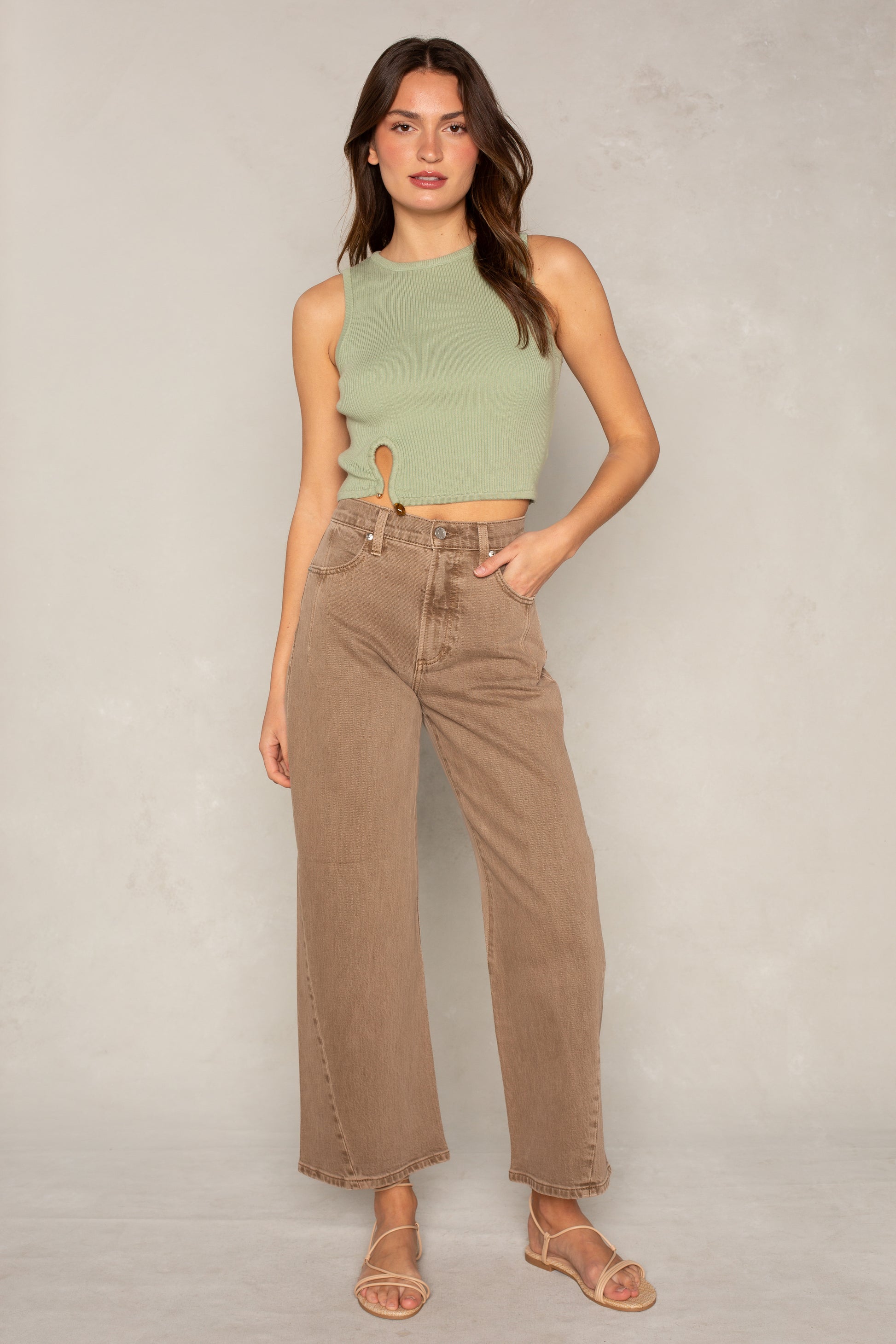 A woman with long brown hair stands against a light grey background wearing the Isla x Tate Tyler Hardware Sweater Tank, high-waisted wide-leg brown pants, and beige sandals. She poses relaxedly with one hand in her pocket.