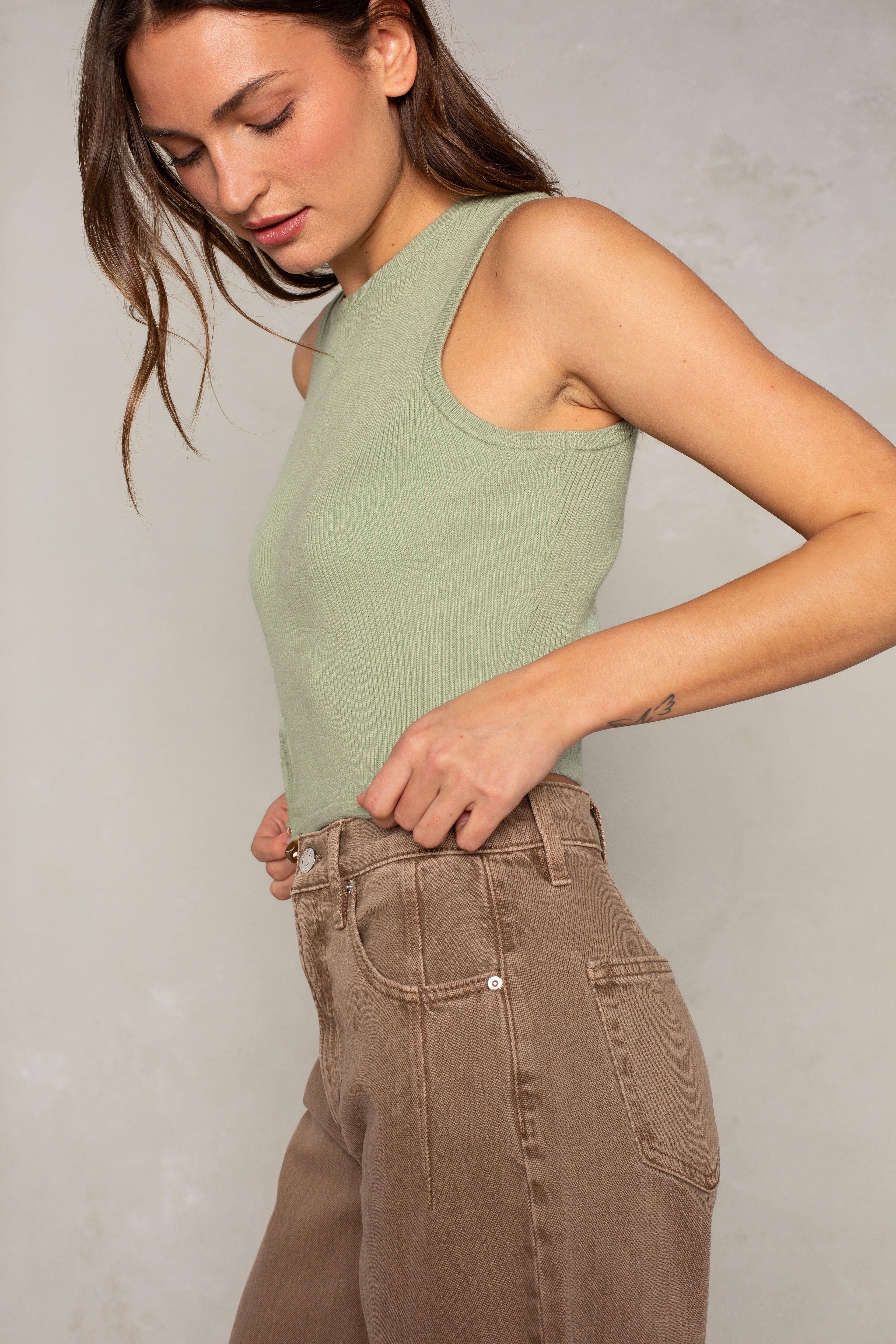 A woman wearing the Isla x Tate Tyler Hardware Sweater Tank, a light green sleeveless ribbed top, and high-waisted brown jeans with asymmetric hardware stands against a neutral background, looking down with one hand on her waistband.