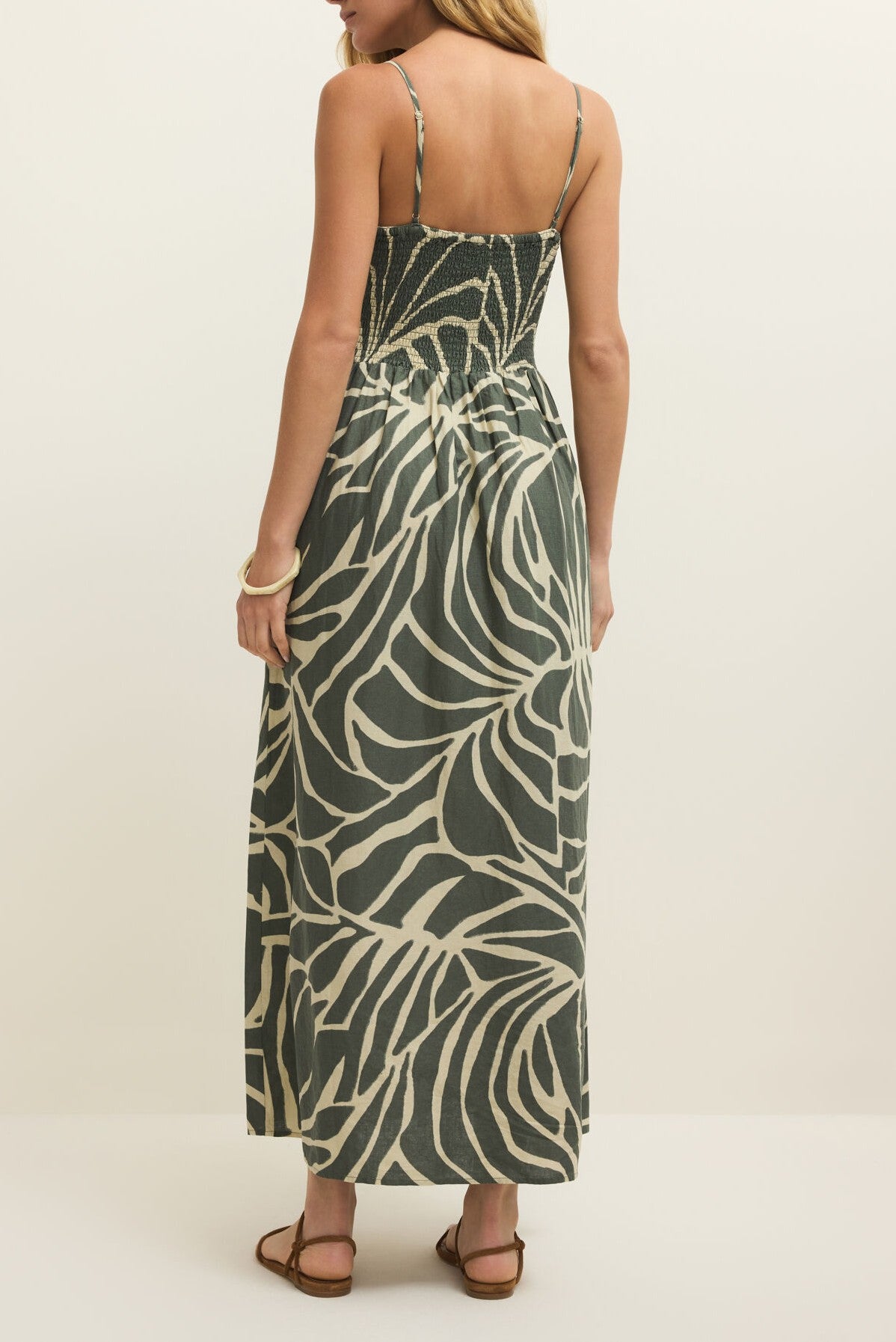 A woman faces away, wearing the Z Supply Bayside Tova Palm Midi Dress with thin straps in dark green and beige, styled with flat brown sandals and a bracelet.