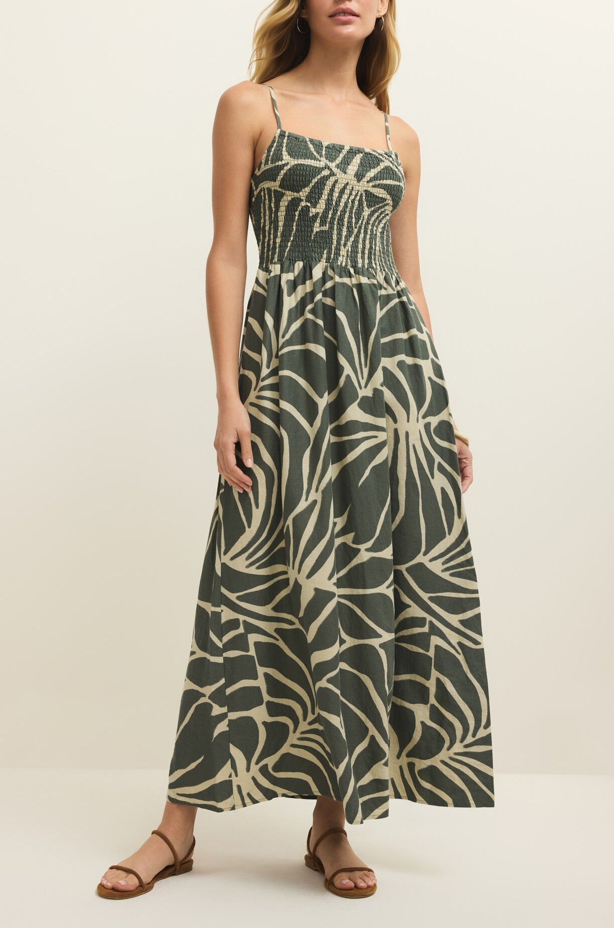 A woman in the Z Supply Bayside Tova Palm Midi Dress—sleeveless, green and beige palm print with smocked bodice and thin straps—paired with brown flat sandals, stands against a plain light background.