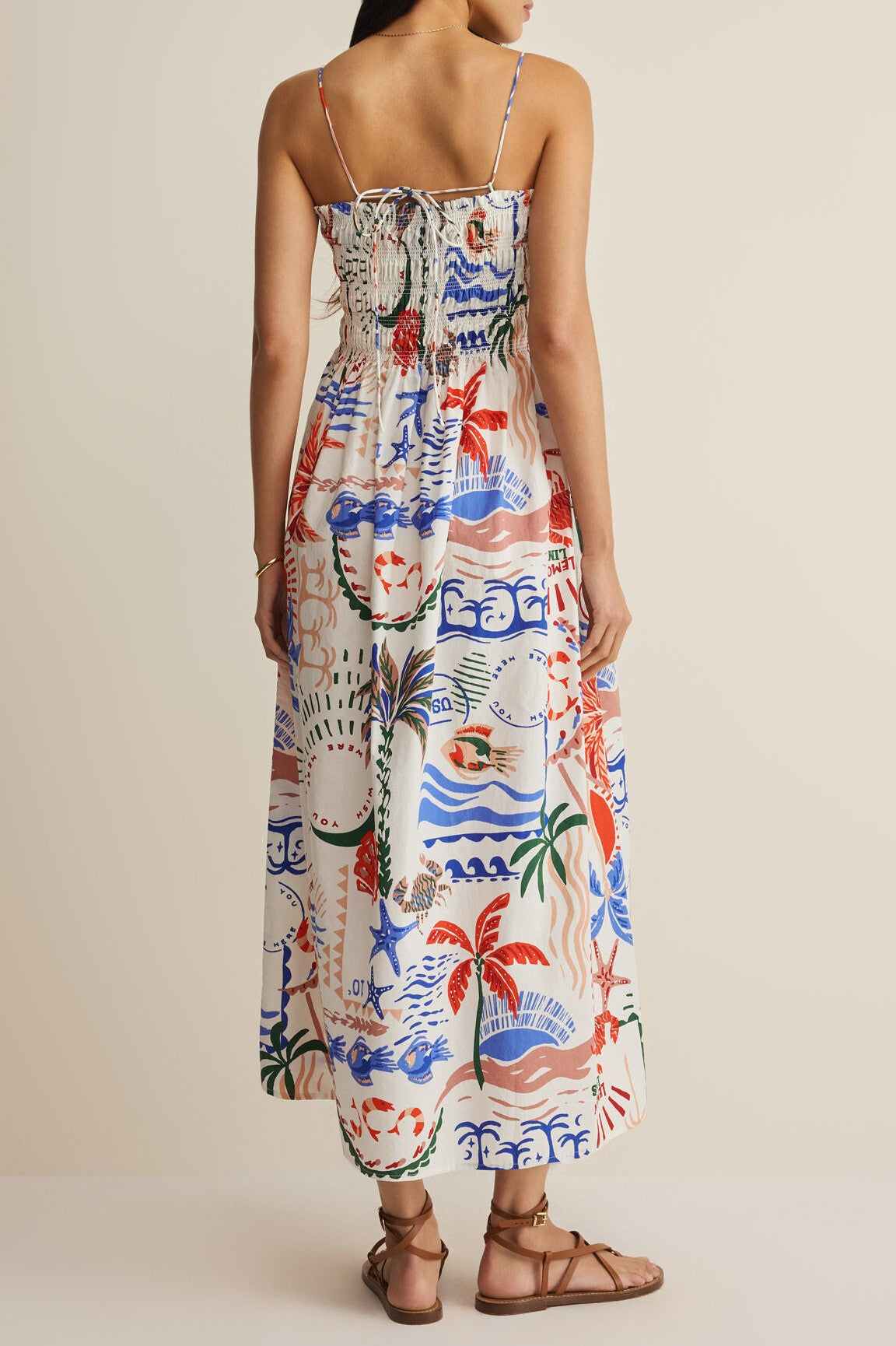 A woman faces away from the camera, wearing the Z Supply Cornelia Getaway Midi Dress with a tropical print, V-neck, spaghetti straps, and side seam pockets, paired with brown flat sandals against a plain beige background.
