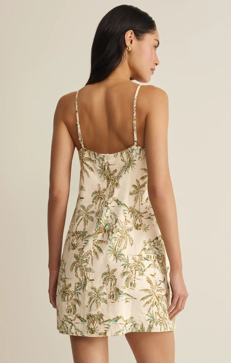 A woman is shown from the back wearing the Maude Tiki Tide Mini Dress by Z Supply—a cream, sleeveless mini with green and brown tropical palm prints, adjustable spaghetti straps, and a fitted silhouette.