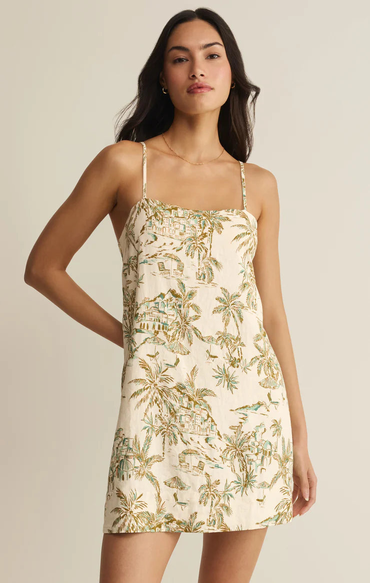 A woman models the Z Supply Maude Tiki Tide Mini Dress in Cream Multi, a washed linen piece with adjustable spaghetti straps and a tropical print, standing confidently against a plain background with one hand on her hip.