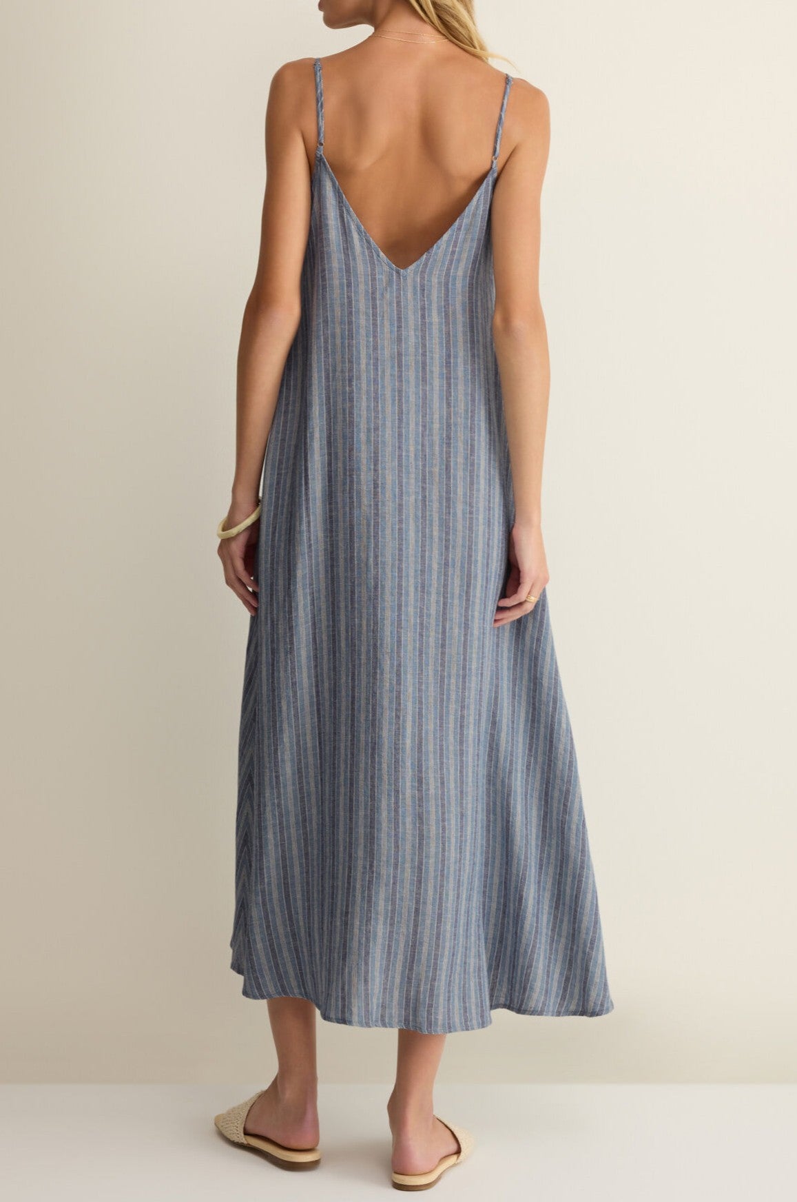 A woman stands facing away in the Z Supply Sandcomber Striped Midi Dress, featuring a sleeveless v-neck, thin straps, and deep V-back. She's barefoot in flat sandals with a bracelet, against a neutral background.