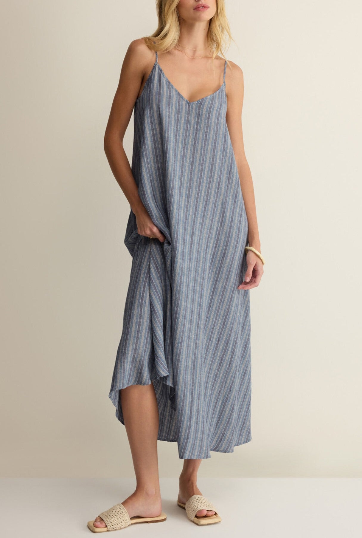 A woman stands against a plain background wearing the Z Supply Sandcomber Striped Midi Dress in light blue, styled with woven beige slide sandals and a light bracelet.