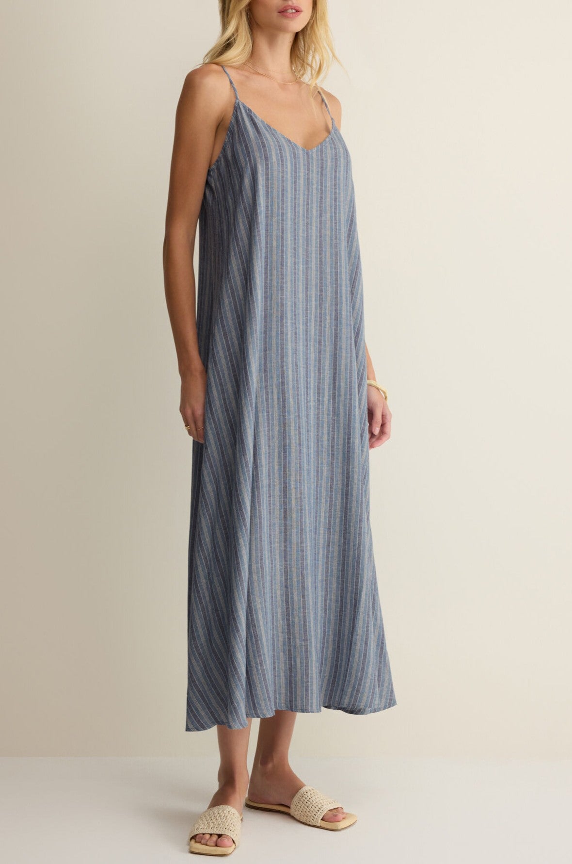 A woman wears the Z Supply Sandcomber Striped Midi Dress—loose, sleeveless, v-neck with thin straps—and beige woven slide sandals, standing against a neutral background.