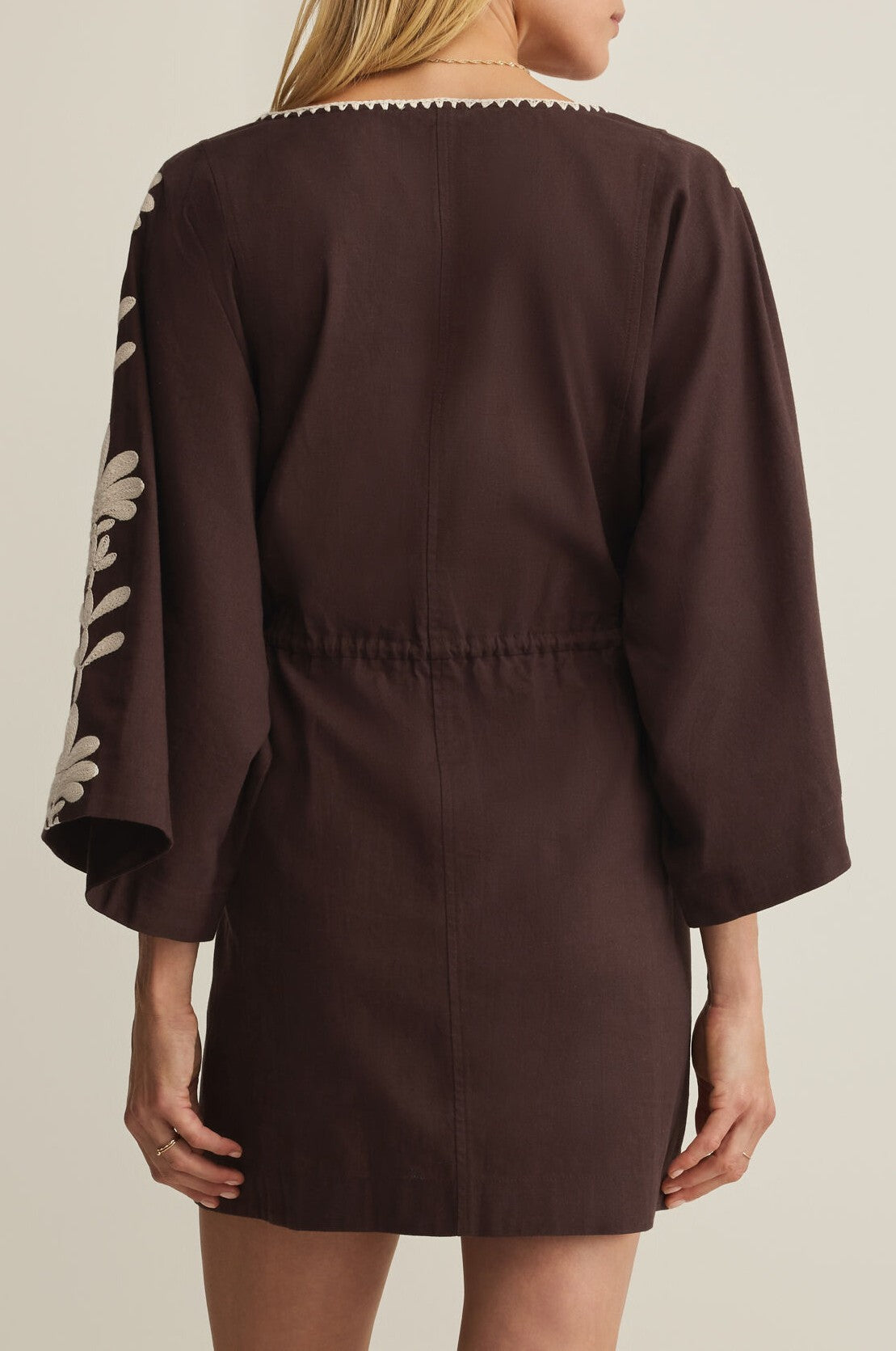 A woman seen from the back wears the Z Supply Whispering Palms Cover Up—a dark brown cotton-linen mini dress featuring a cinched waist, relaxed fit, and white embroidered floral detail on one sleeve.