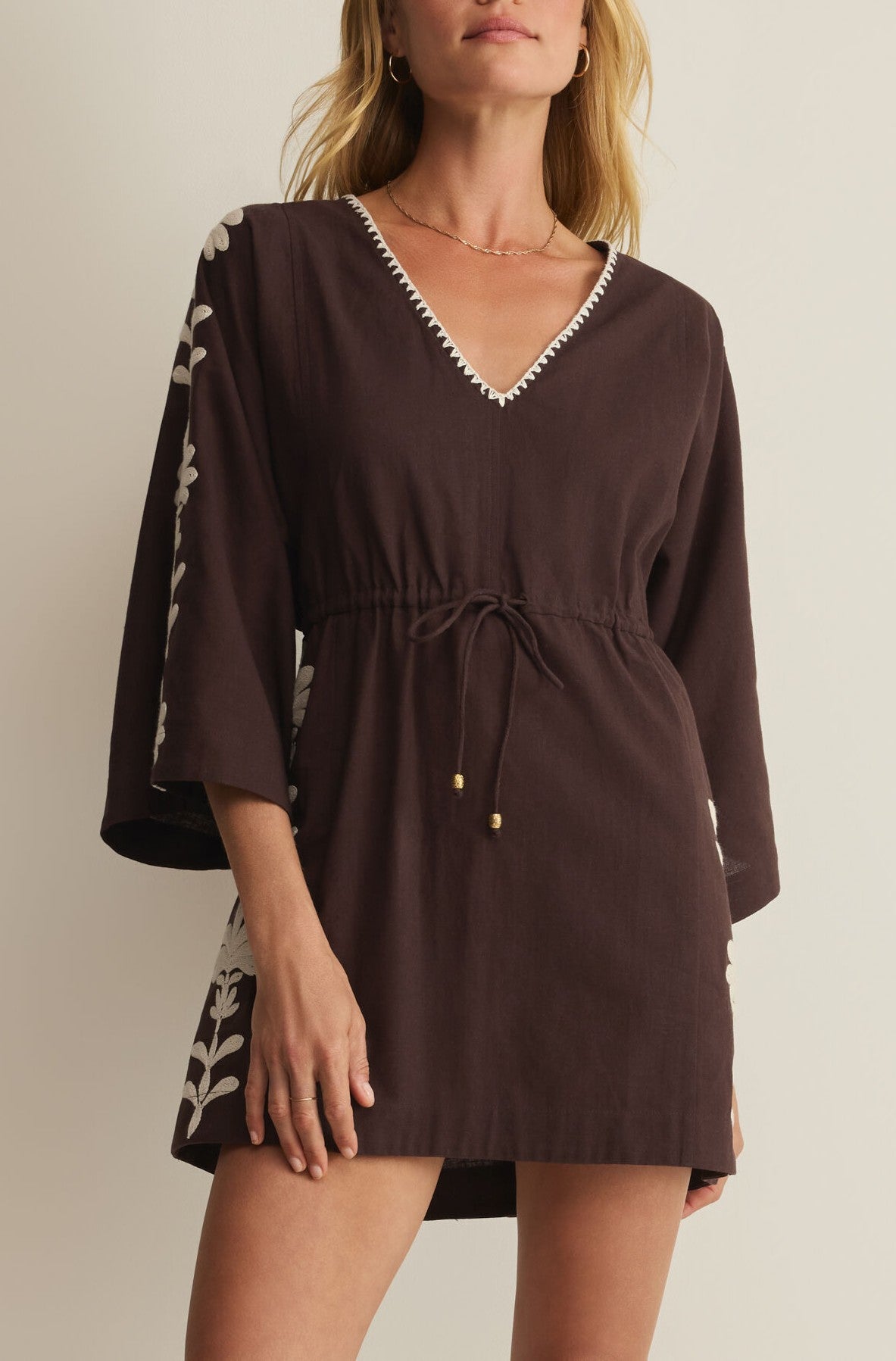 A woman wears the Z Supply Whispering Palms Cover Up in brown, featuring a V-neck mini silhouette with white floral embroidery on the sleeves and sides, an elastic waistband for comfort, and subtle neckline trim.