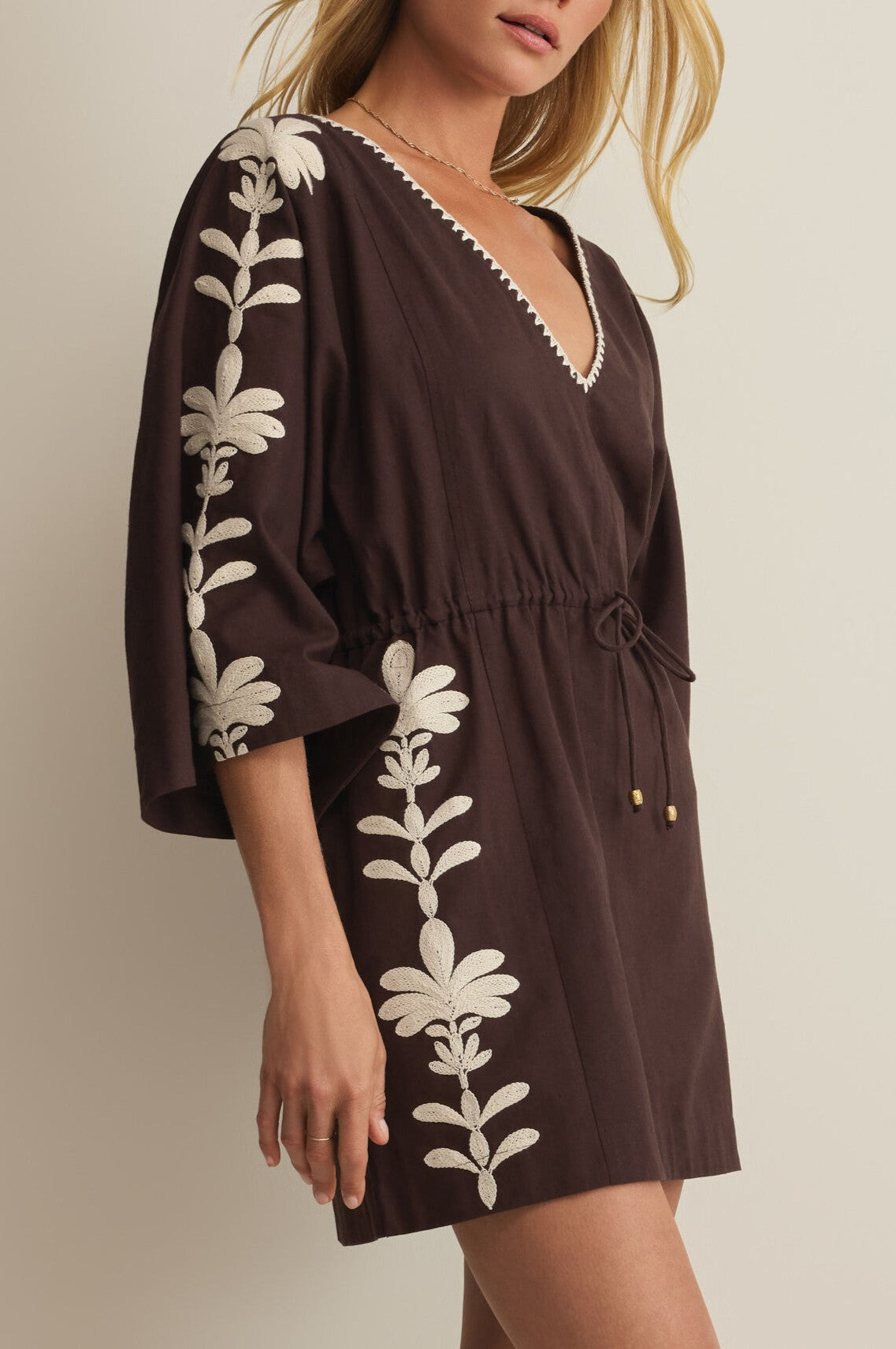 A woman wears the Whispering Palms Cover Up by Z Supply, a dark brown cotton-linen mini dress with loose sleeves, cream embroidered leaf accents, and a drawstring cinched waist for a flattering fit.