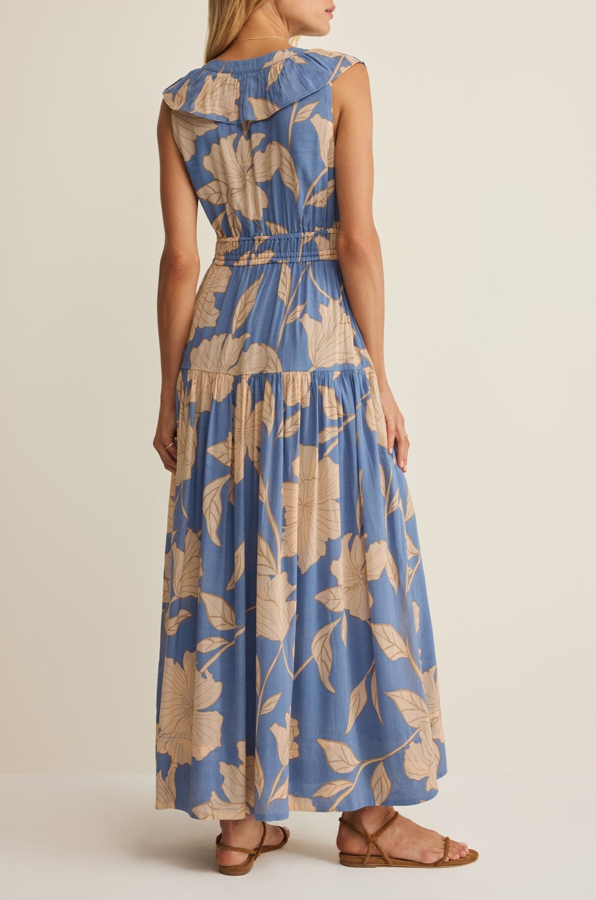 A woman faces away, wearing the Z Supply Malene Bloom Floral Maxi Dress—a sleeveless cotton voile sundress with beige floral prints, a tiered skirt, gathered waist, and wide collar—paired with brown flat sandals.