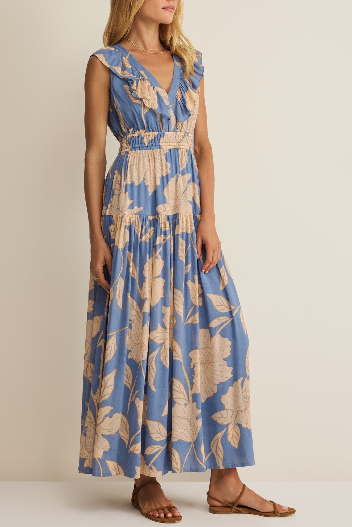 A woman wears the Z Supply Malene Bloom Floral Maxi Dress—sleeveless, blue with beige floral print, ruffled shoulders, V-neckline, elastic waist, and tiered skirt—paired with flat brown sandals against a plain background.