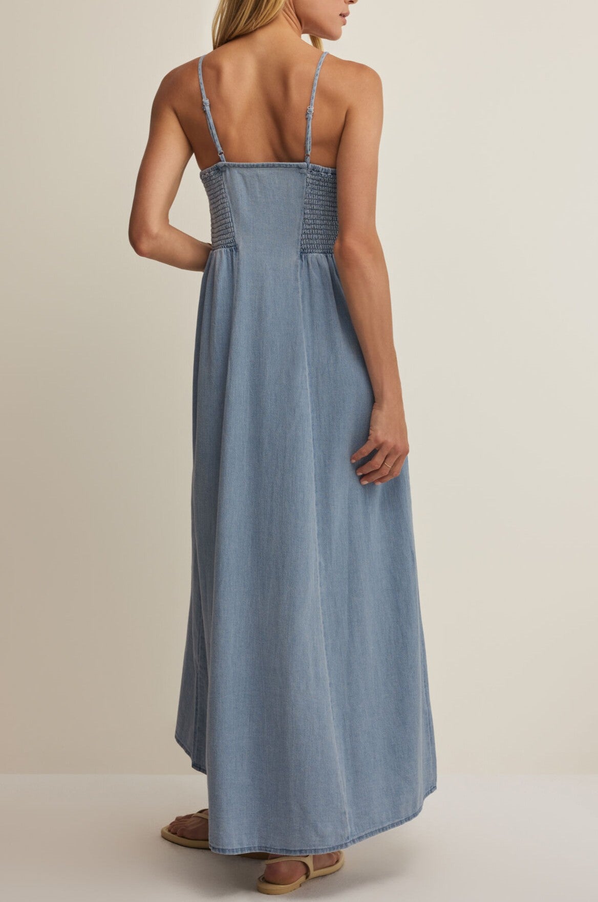 A woman wearing the Angel Eyes Twill Midi Dress by Z Supply, featuring a light blue cotton fabric with thin straps and a smocked back, stands facing away from the camera against a plain background, paired with flat sandals.