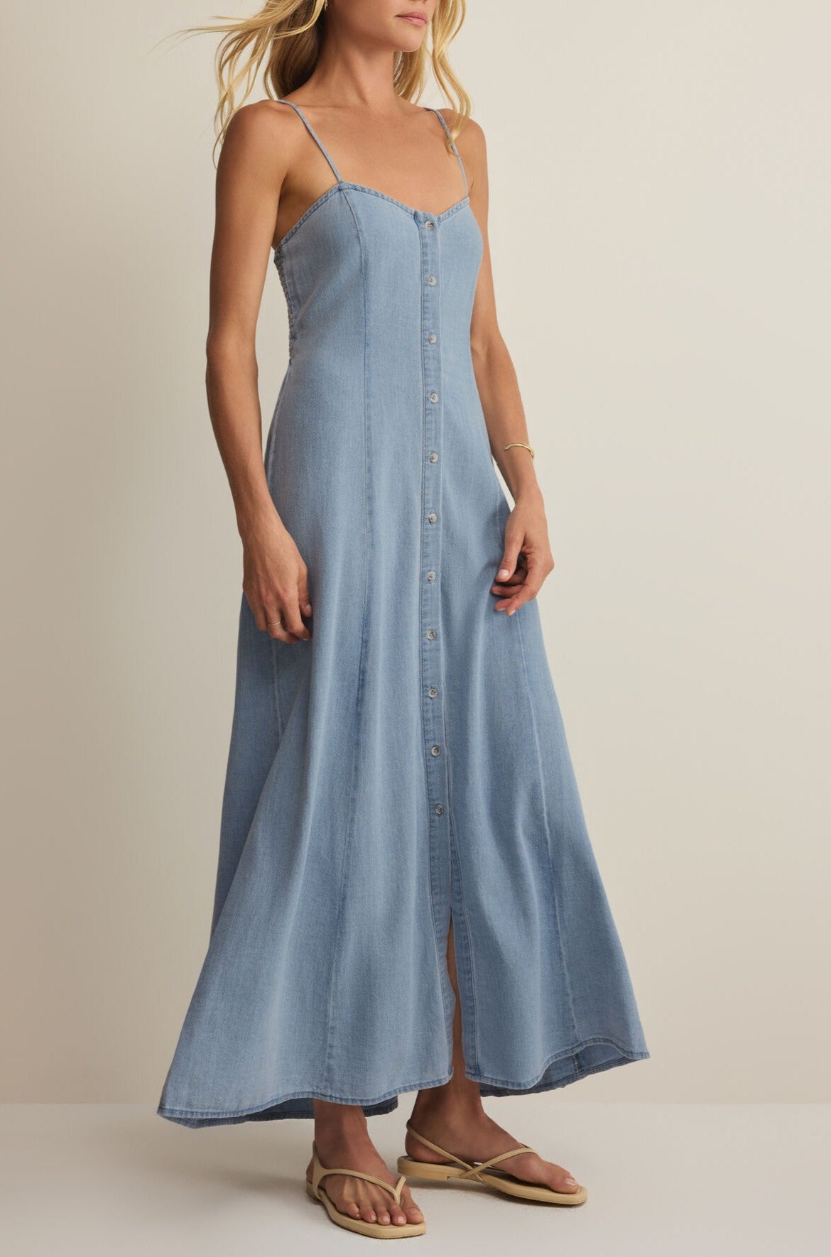A woman in the Z Supply Angel Eyes Twill Midi Dress, light blue with thin straps, and beige flip-flops stands against a neutral background.