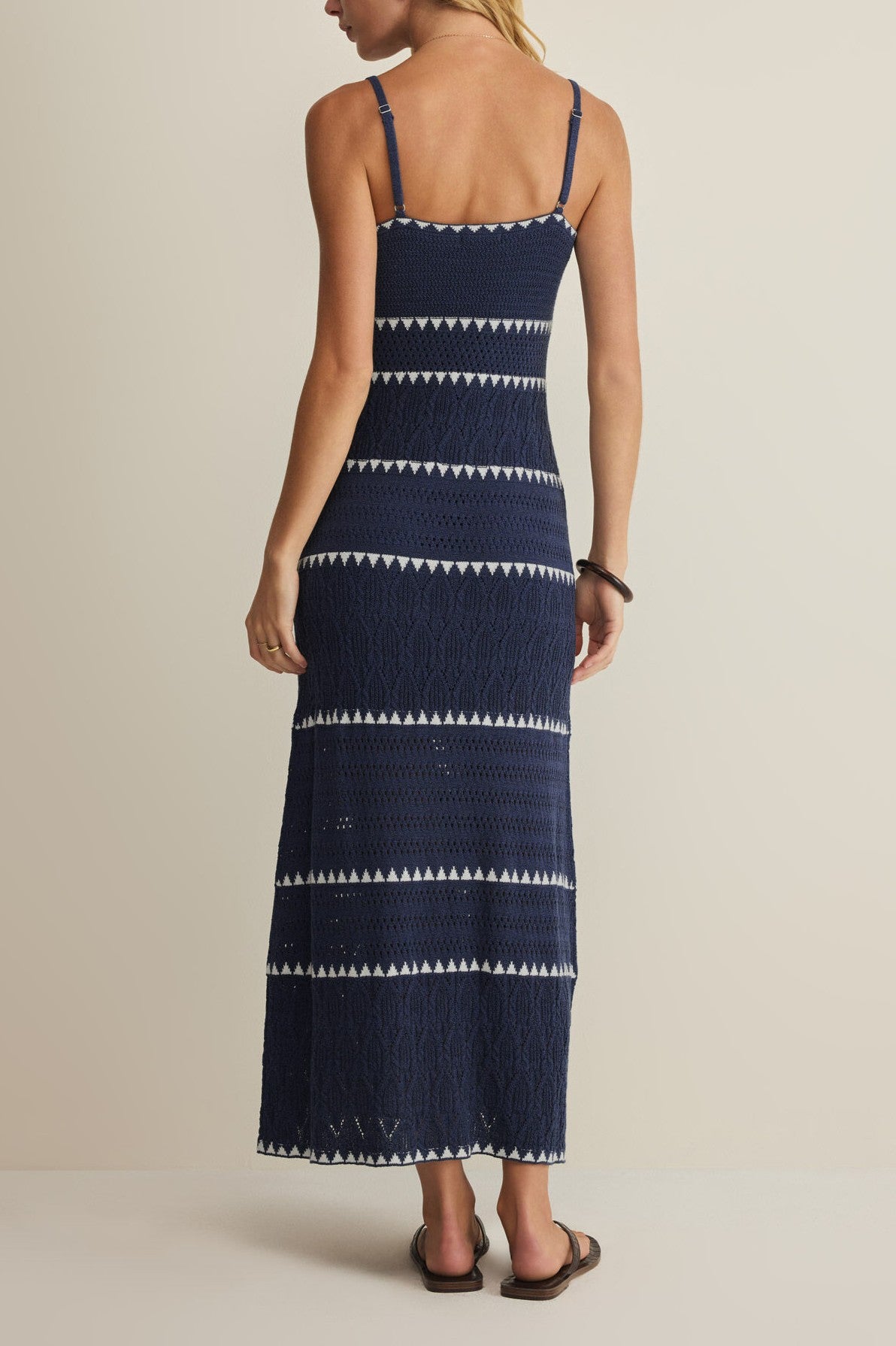 A woman faces away, wearing the Z Supply Catamaran Striped Maxi Dress in navy with white geometric stripes and black sandals, set against a plain light beige background.