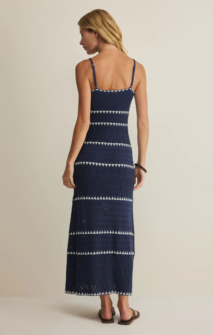 A woman with long blonde hair stands facing away, wearing the Z Supply Catamaran Striped Maxi Dress—navy blue and white, sleeveless with a scoop neck and thin straps—paired with black sandals against a plain light background.