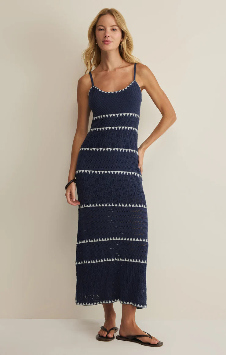 A woman stands against a plain light wall wearing the Catamaran Striped Maxi Dress by Z Supply—navy blue, sweater knit, sleeveless with thin straps and white zigzag stripes—paired with black sandals, one hand on her hip.