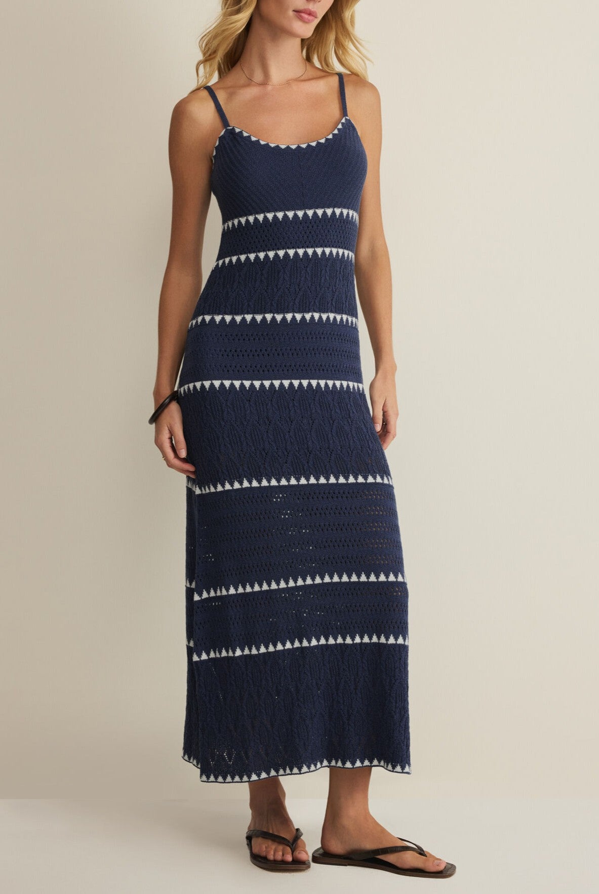 A woman wears the Z Supply Catamaran Striped Maxi Dress—navy blue, sleeveless with white geometric stripes and spaghetti straps—paired with black flip-flops, standing against a plain light background.