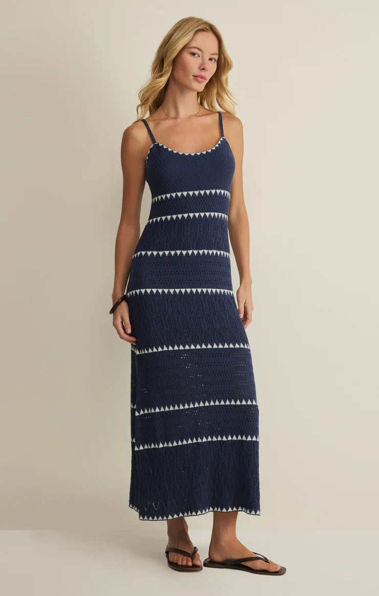 A woman with blonde hair and a relaxed expression wears the Z Supply Catamaran Striped Maxi Dress, a navy blue, sleeveless maxi dress with white geometric stripes, against a light background. She pairs it with black flip-flops.