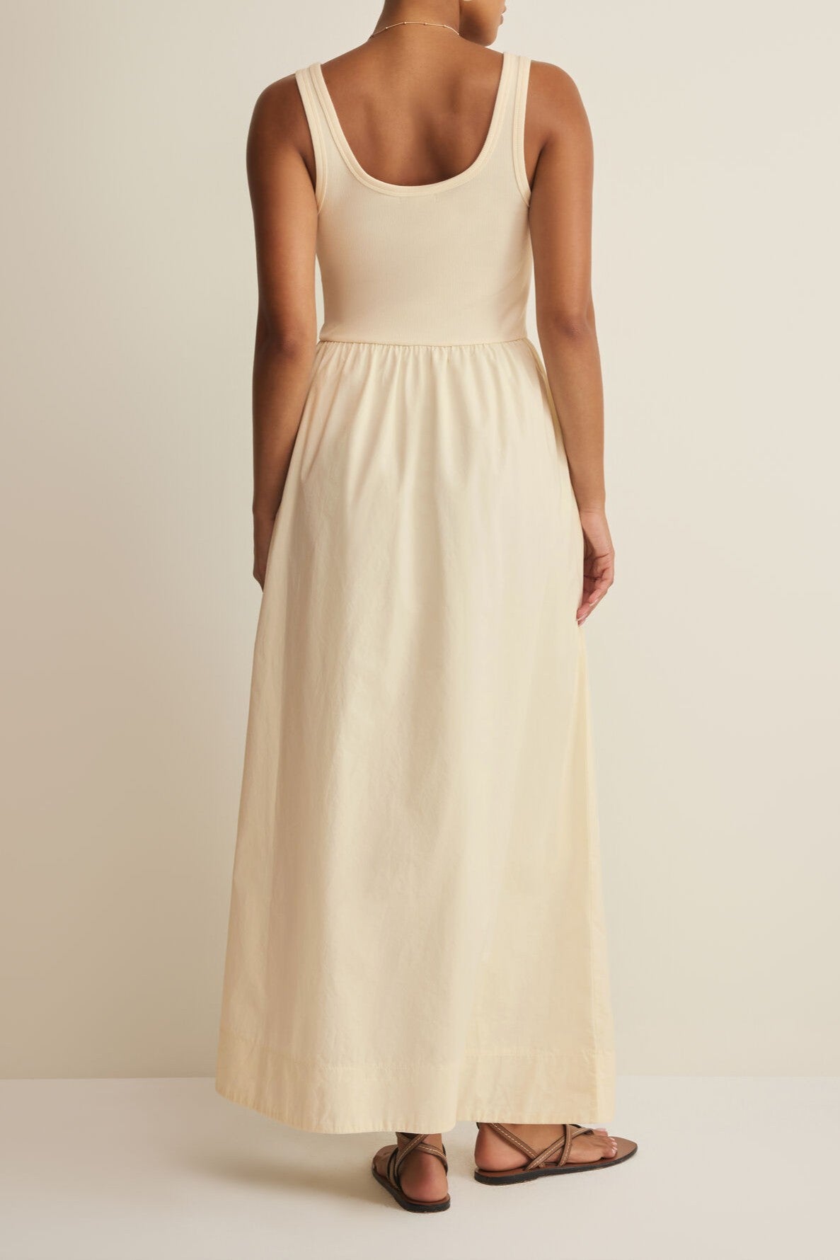 A person faces away, wearing the Z Supply Kensie Midi Dress—a sleeveless, light cream midi with a fitted top and gathered cotton skirt—paired with brown sandals against a plain light background.
