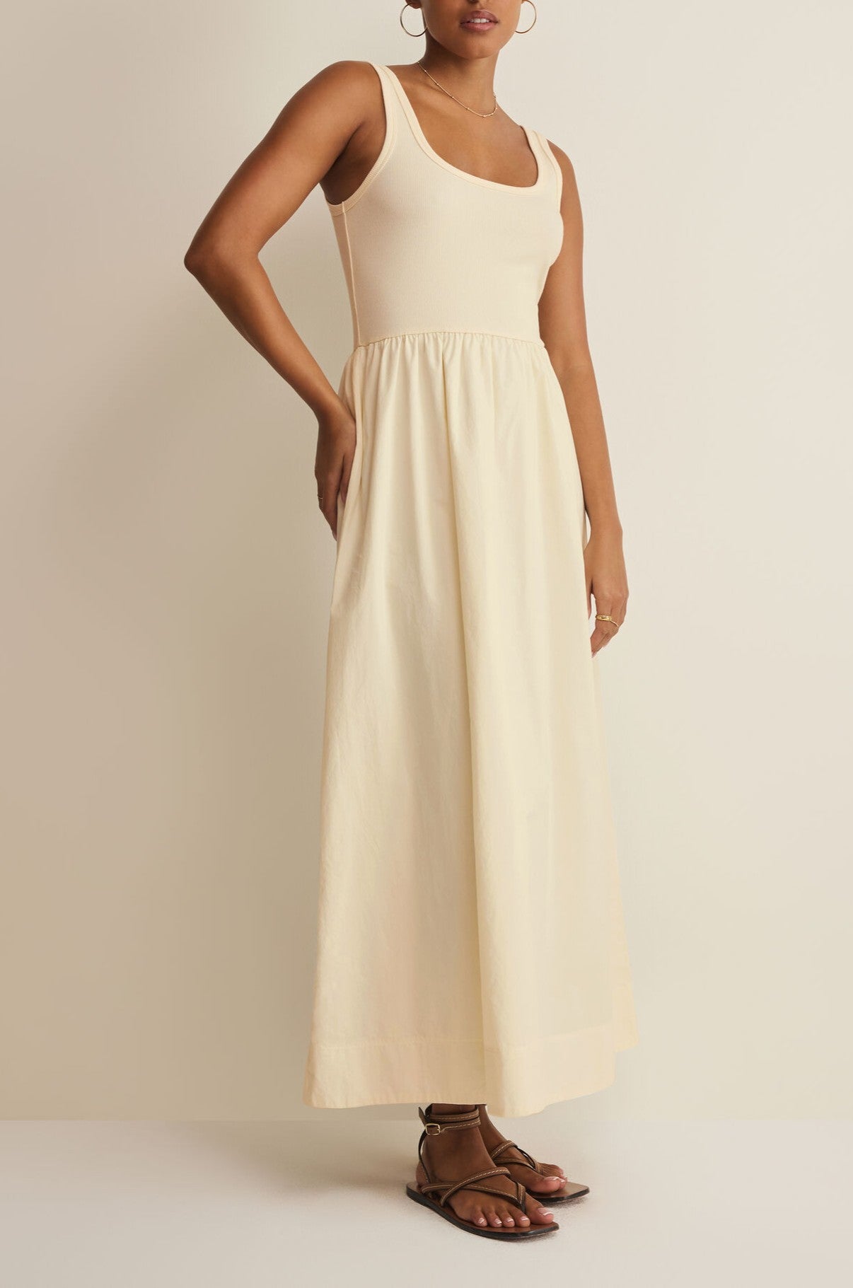 A woman wears the Kensie Midi Dress by Z Supply, a sleeveless cream ribbed dress with a fitted bodice and flowing skirt, paired with brown strappy sandals, standing against a plain light background.