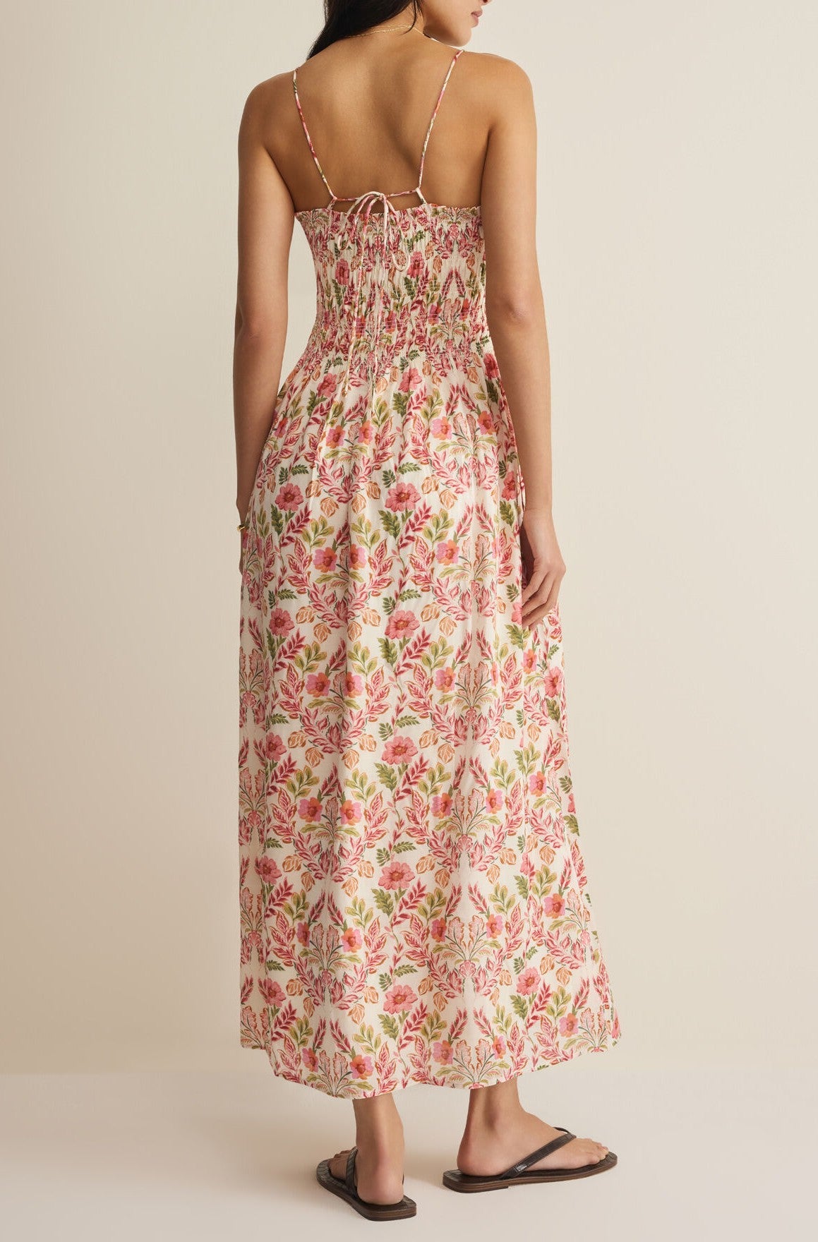 A woman faces away wearing the Z Supply Cornelia Ami Floral Midi Dress, a sleeveless cotton-rayon style with thin straps, pink and green florals, a smocked bodice, and black sandals. The background is plain and neutral.