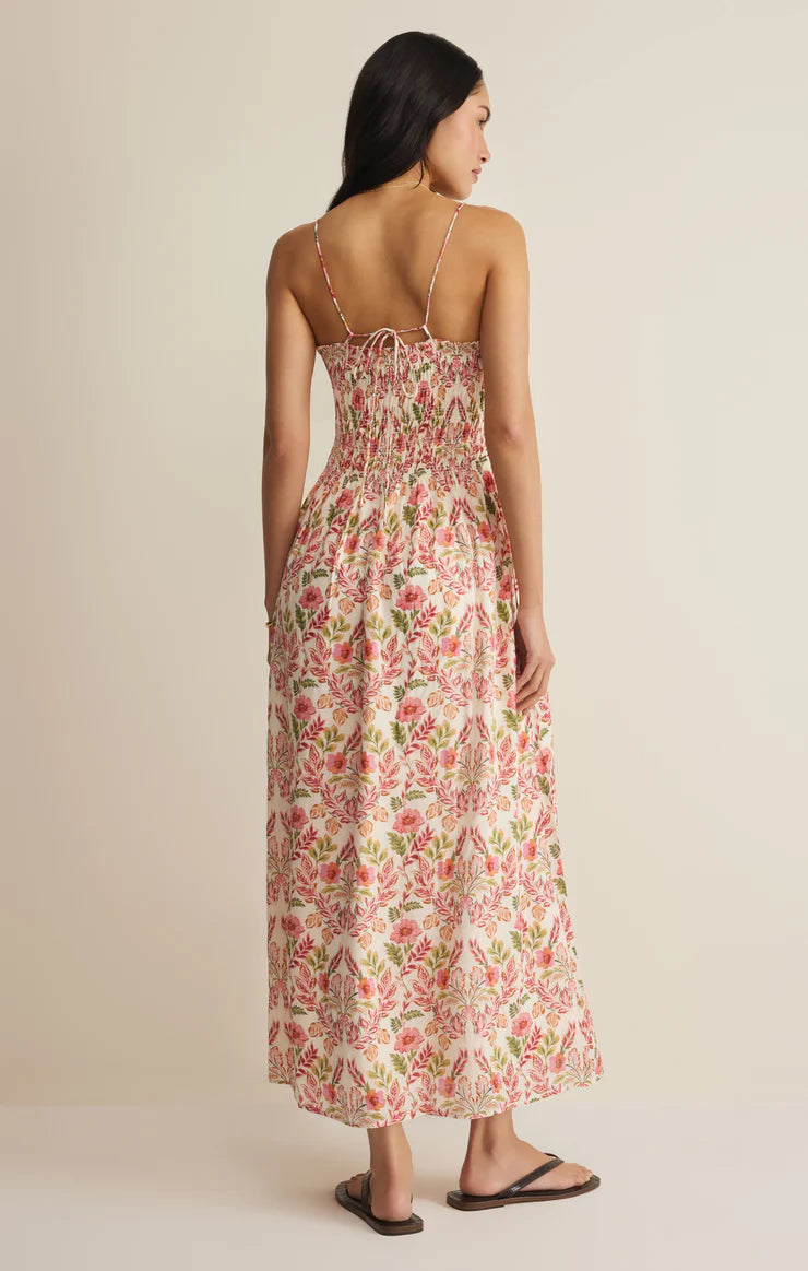 A woman with long dark hair faces away, wearing the Z Supply Cornelia Ami Floral Midi Dress with adjustable spaghetti straps and a pink and green floral print. She pairs it with black sandals against a neutral background.