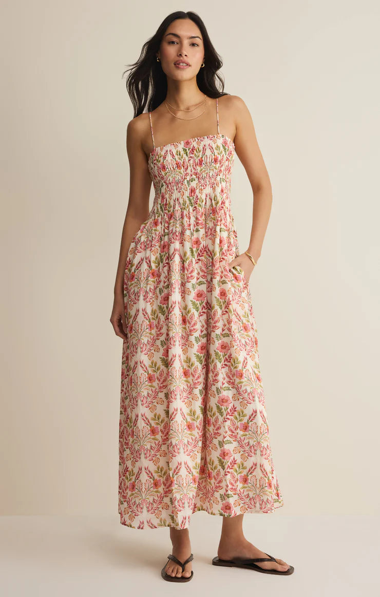A woman with long dark hair wears the Z Supply Cornelia Ami Floral Midi Dress in Pink Multi—featuring adjustable spaghetti straps, a smocked bodice, and floral print—paired with sandals against a neutral backdrop.