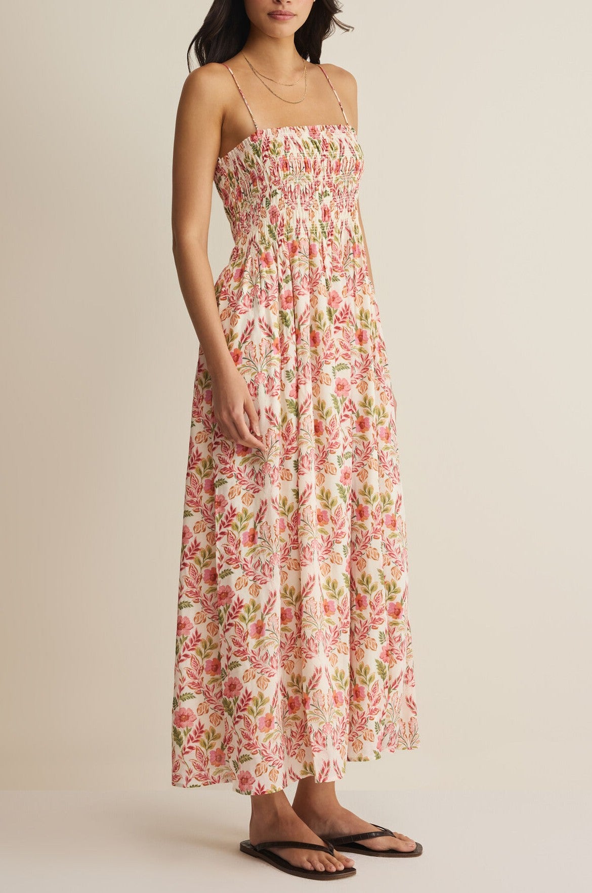 A woman wears the Z Supply Cornelia Ami Floral Midi Dress, a sleeveless cotton-rayon dress with thin straps, smocked bodice, pink and green floral pattern on a light background. She pairs it with black sandals.