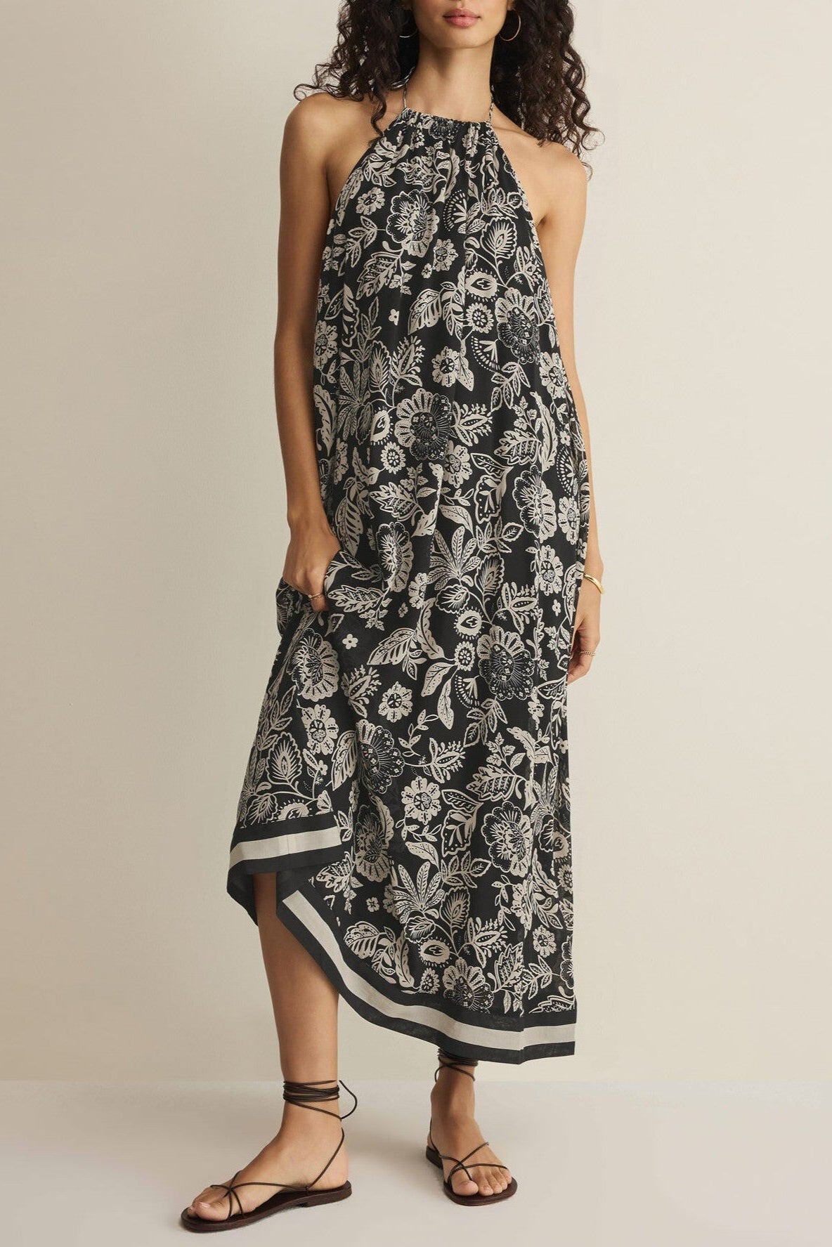 A woman stands against a plain light background wearing the Viento Luz Floral Midi Dress by Z Supply, a sleeveless halter-neck summer dress with a black and white floral print, paired with strappy black sandals.