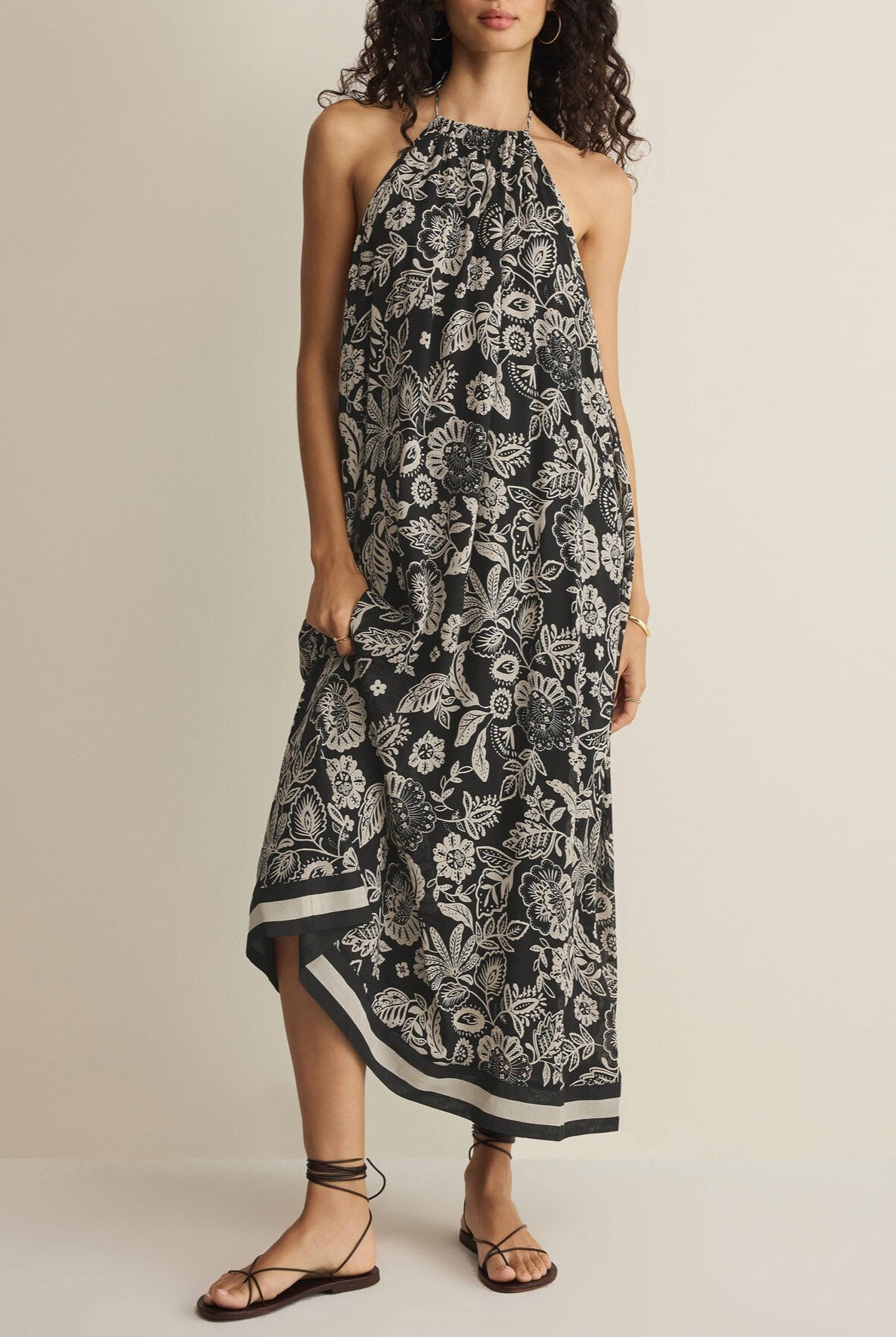 A woman stands against a plain light background wearing the Viento Luz Floral Midi Dress by Z Supply, a sleeveless halter-neck summer dress with a black and white floral print, paired with strappy black sandals.