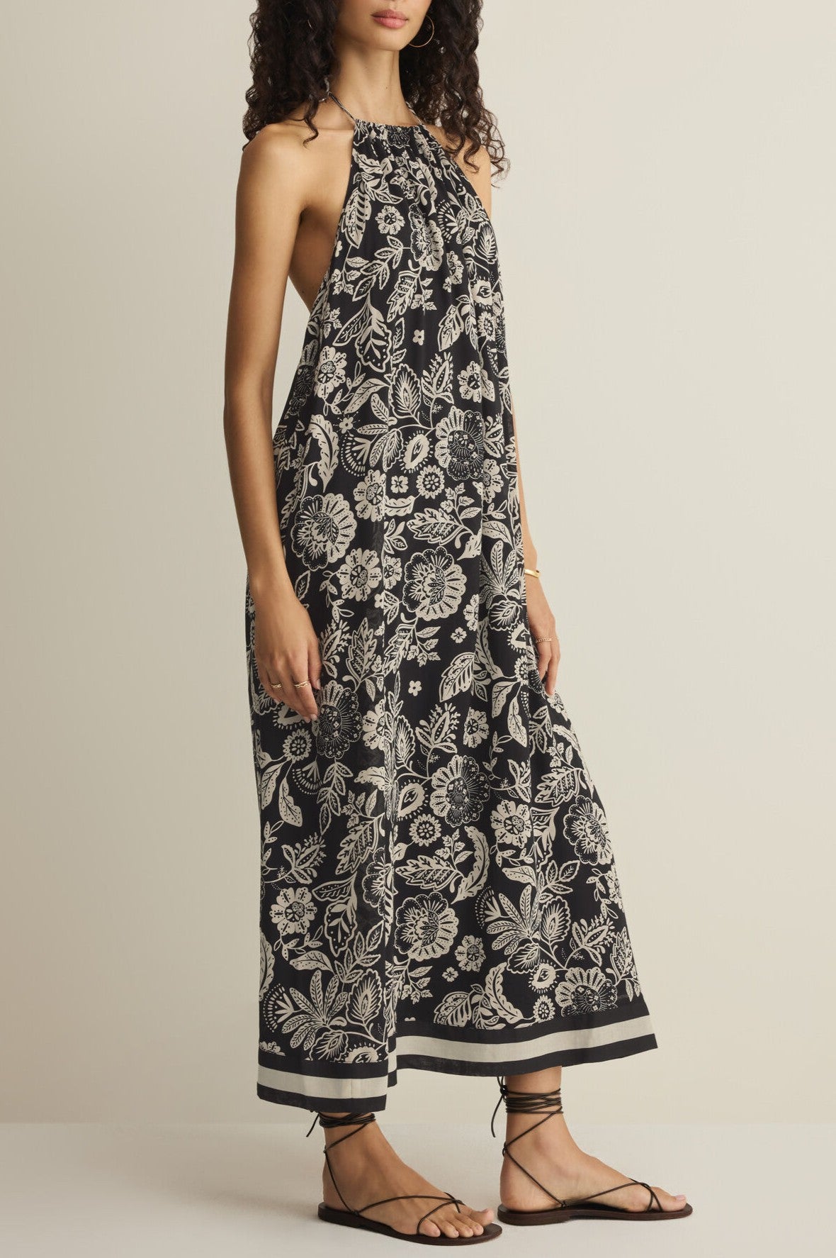 Model wears the Z Supply Viento Luz Floral Midi Dress— a sleeveless, halter-neck black sundress with white floral print—paired with black strappy sandals, standing against a plain background.