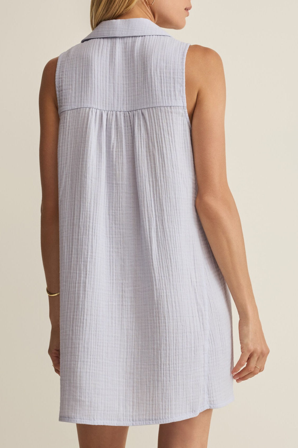 A woman is seen from behind in the Z Supply New Light Mini Dress—a collared, sleeveless, light gray cotton dress with back yoke pleats. She accessorizes with a gold bracelet on her right wrist.