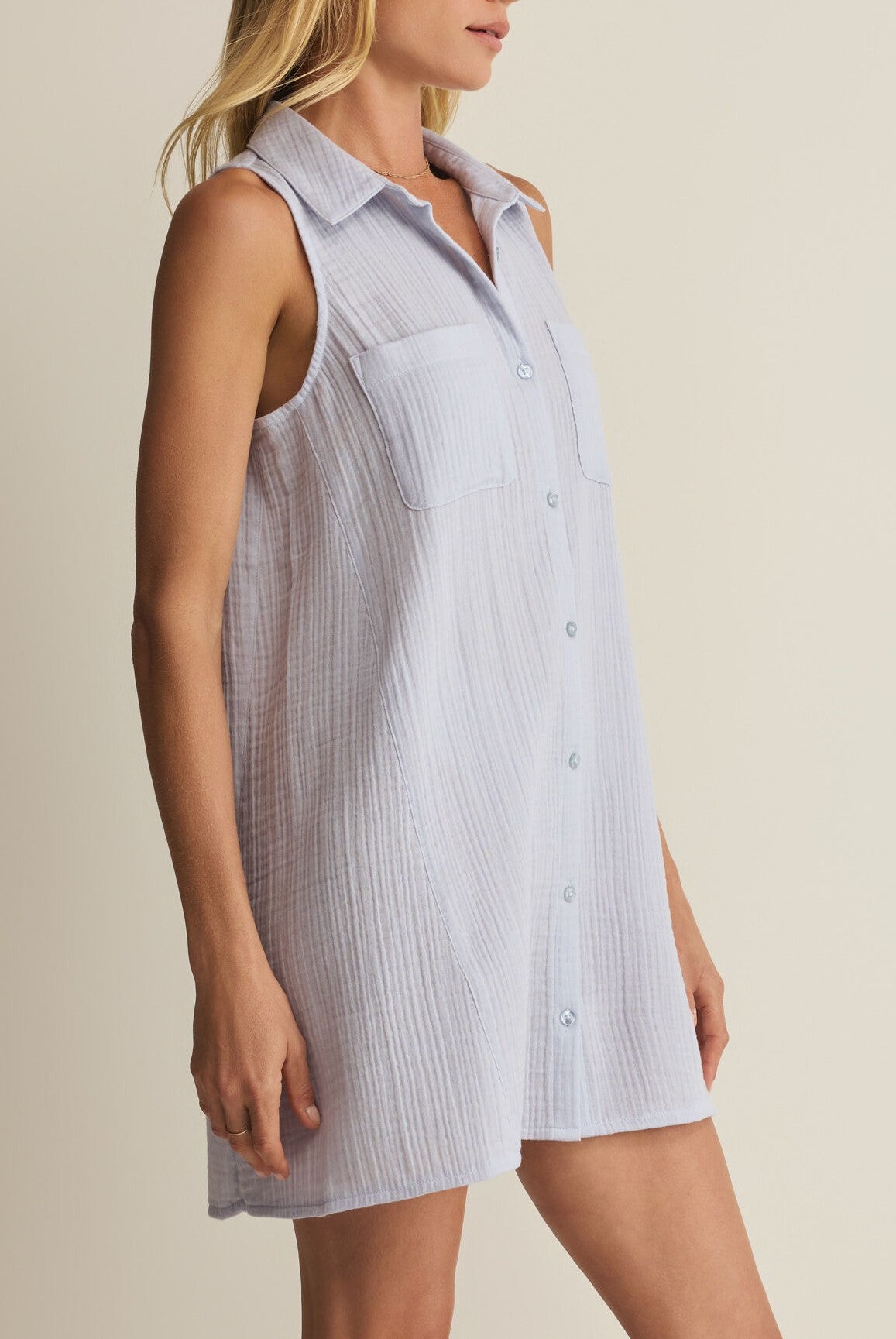A woman wears the Z Supply New Light Mini Dress, a collared sleeveless mini dress in light blue textured cotton with two chest pockets and a relaxed fit.