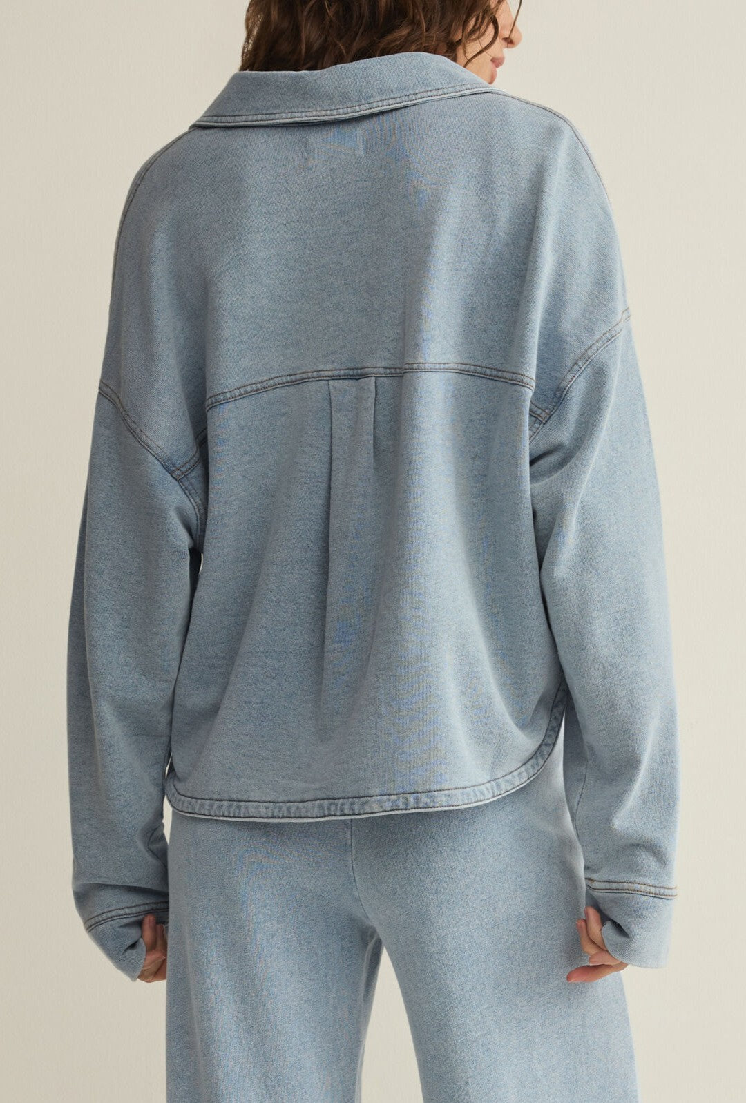 A person with wavy brown hair is shown from the back, wearing the Z Supply All Day Cropped Knit Jacket in light blue, featuring a collar, long sleeves, pleat and seam detail across the upper back, paired with matching pants.
