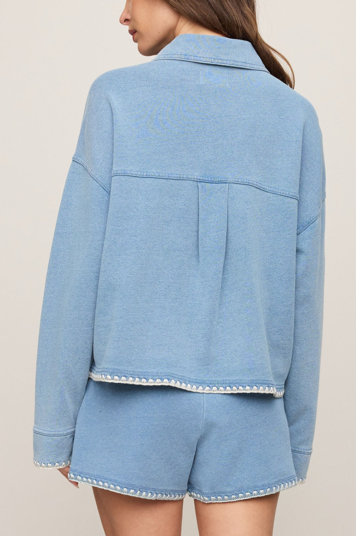 A woman is seen from the back wearing Z Supply's All Day Crop Whipstitch Jacket in light blue knit denim with matching frayed-hem shorts, set against a plain beige background.