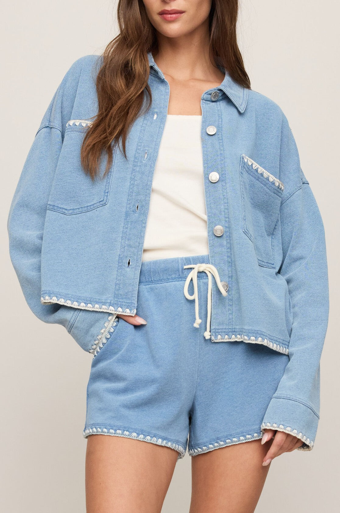 A woman wears the Z Supply All Day Crop Whipstitch Jacket in blue, styled with matching drawstring shorts and a white tank top, standing against a neutral background with one hand in her pocket.