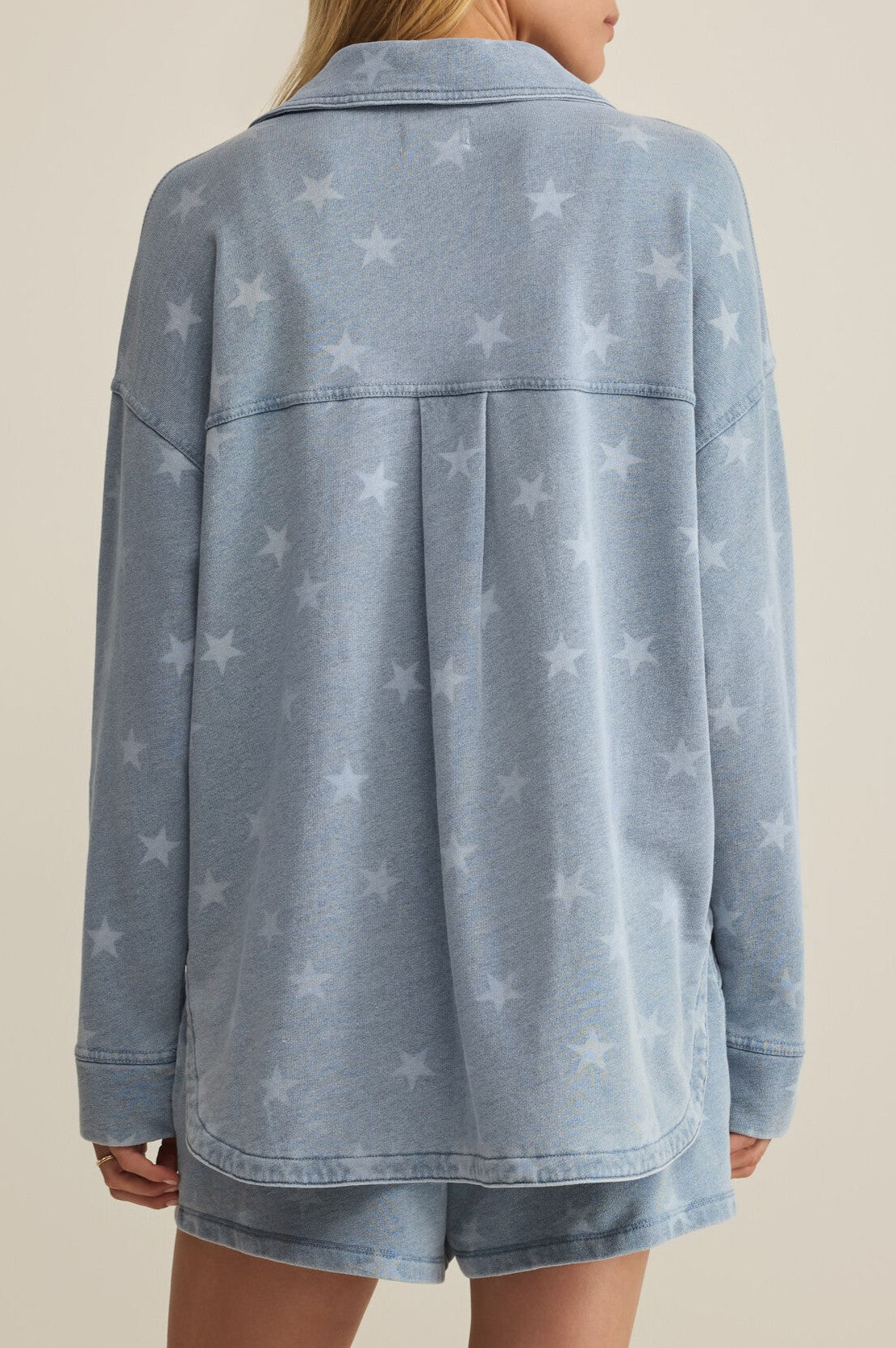 A person wears the Z Supply All Day Knit Star Jacket in light blue, featuring long sleeves and a subtle star pattern, paired with matching shorts. The photo displays the back view of the outfit against a plain, light background.