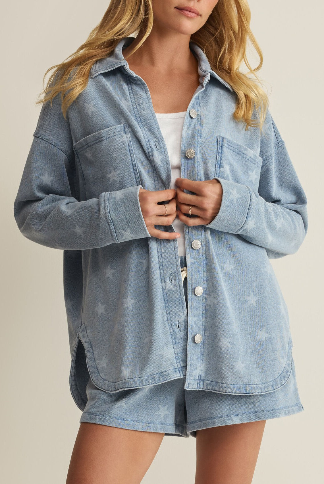 A woman with long blonde hair wears the Z Supply All Day Knit Star Jacket in light blue over a white top and matching shorts, standing with hands near the buttons against a neutral background.