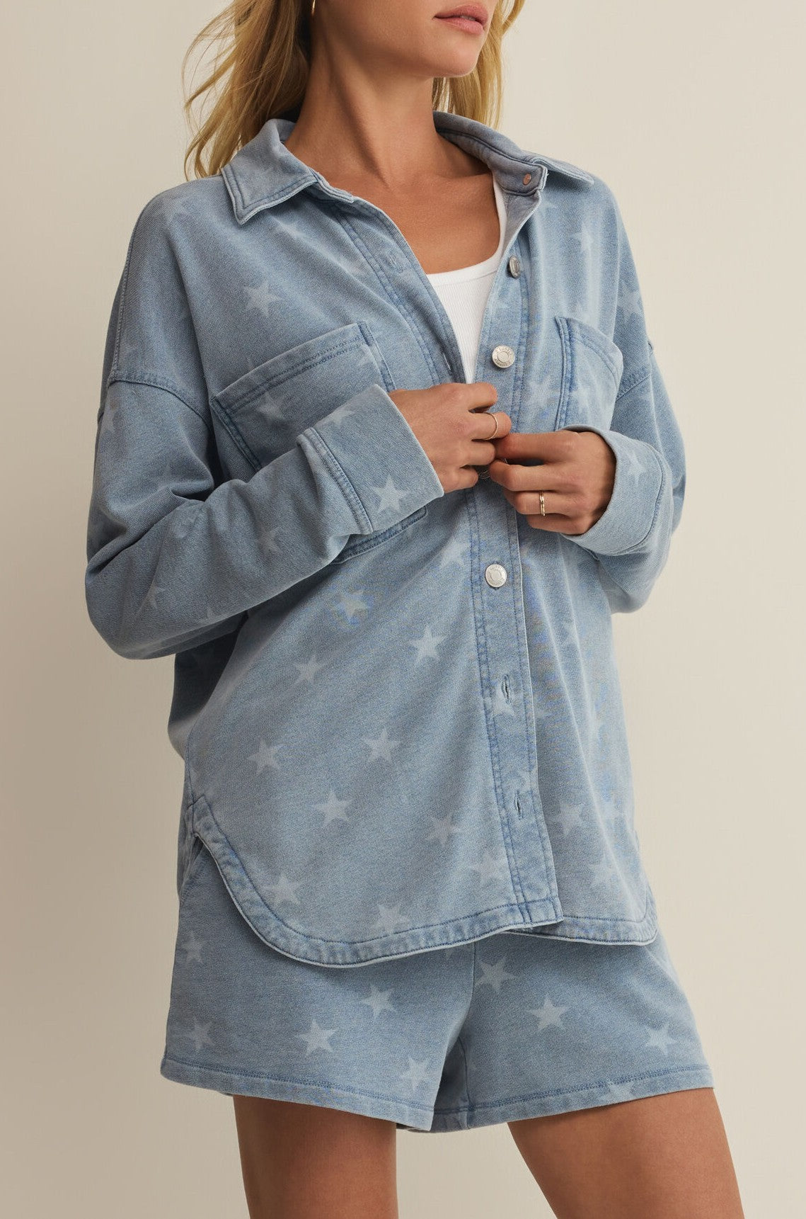 A woman stands against a neutral background wearing the Z Supply All Day Knit Star Jacket—a light blue, star-patterned denim shirt jacket—over a white top with matching shorts.