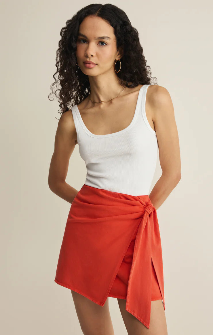 A woman with long curly hair wears a white cotton jersey tank top and the Z Supply Sora Mini Skirt in bright orange. She stands against a beige background, looking at the camera with a neutral expression.