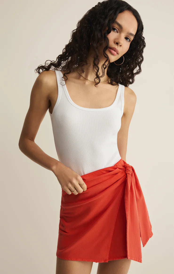 A woman with long, curly hair models the Z Supply Sora Mini Skirt, a red high-rise wrap style with a waist knot, paired with a white sleeveless cotton jersey tank top, posing against a neutral background.