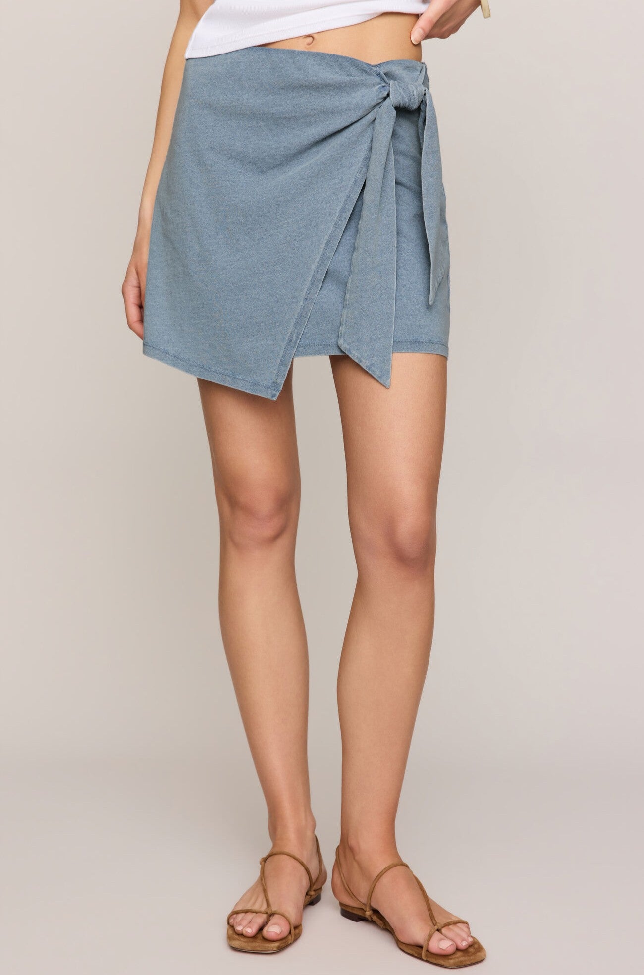A woman wears the Z Supply Sorajersey Denim Mini Skirt in light blue, paired with a white top and brown flat sandals, standing against a neutral background.