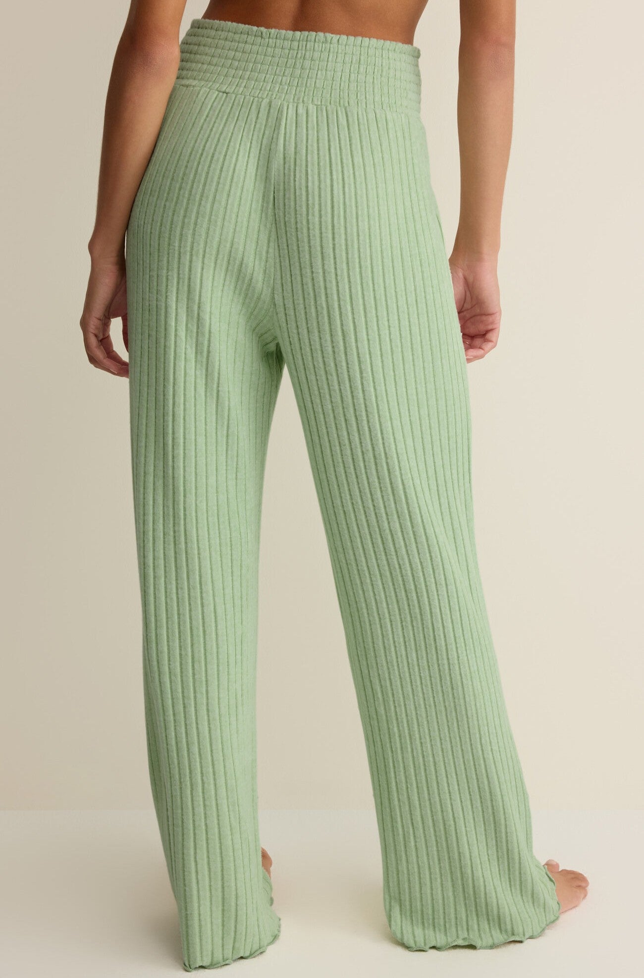 A person is shown from the back wearing Z Supply’s Dawn Smocked Rib Pant in light green. These high-rise, loose-fitting ribbed knit pants feature a wide-leg design and smocked waistband, reaching down to the ankles against a plain neutral background.