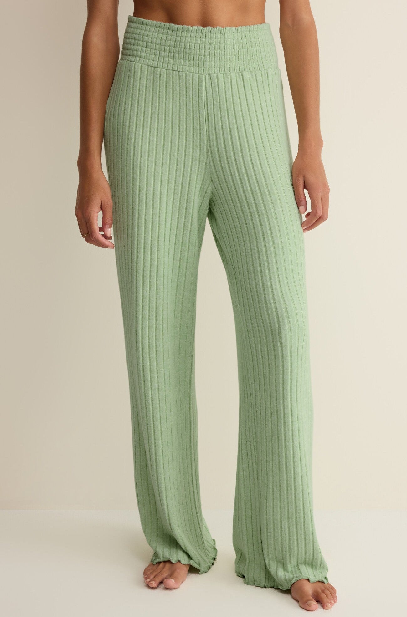 A person stands barefoot in the Z Supply Dawn Smocked Rib Pant, light green ribbed knit with a smocked waistband and relaxed, floor-length fit. Crafted from silky rib fabric, the scene is set against a neutral beige background.