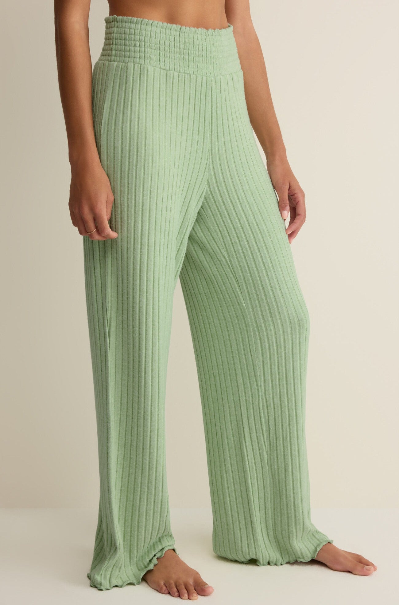 A person stands barefoot against a neutral background wearing the Z Supply Dawn Smocked Rib Pant, featuring a high rise, light green hue, ribbed knit fabric, and a smocked waistband.